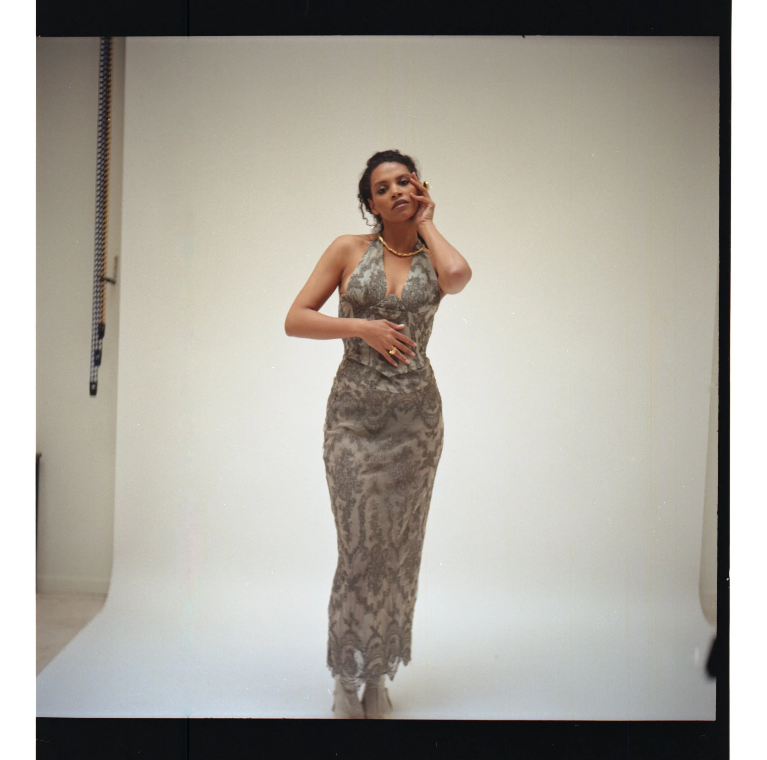 Femme portant une robe longue en tissu avec un motif complexe, posant devant un fond blanc dans un studio photographique.