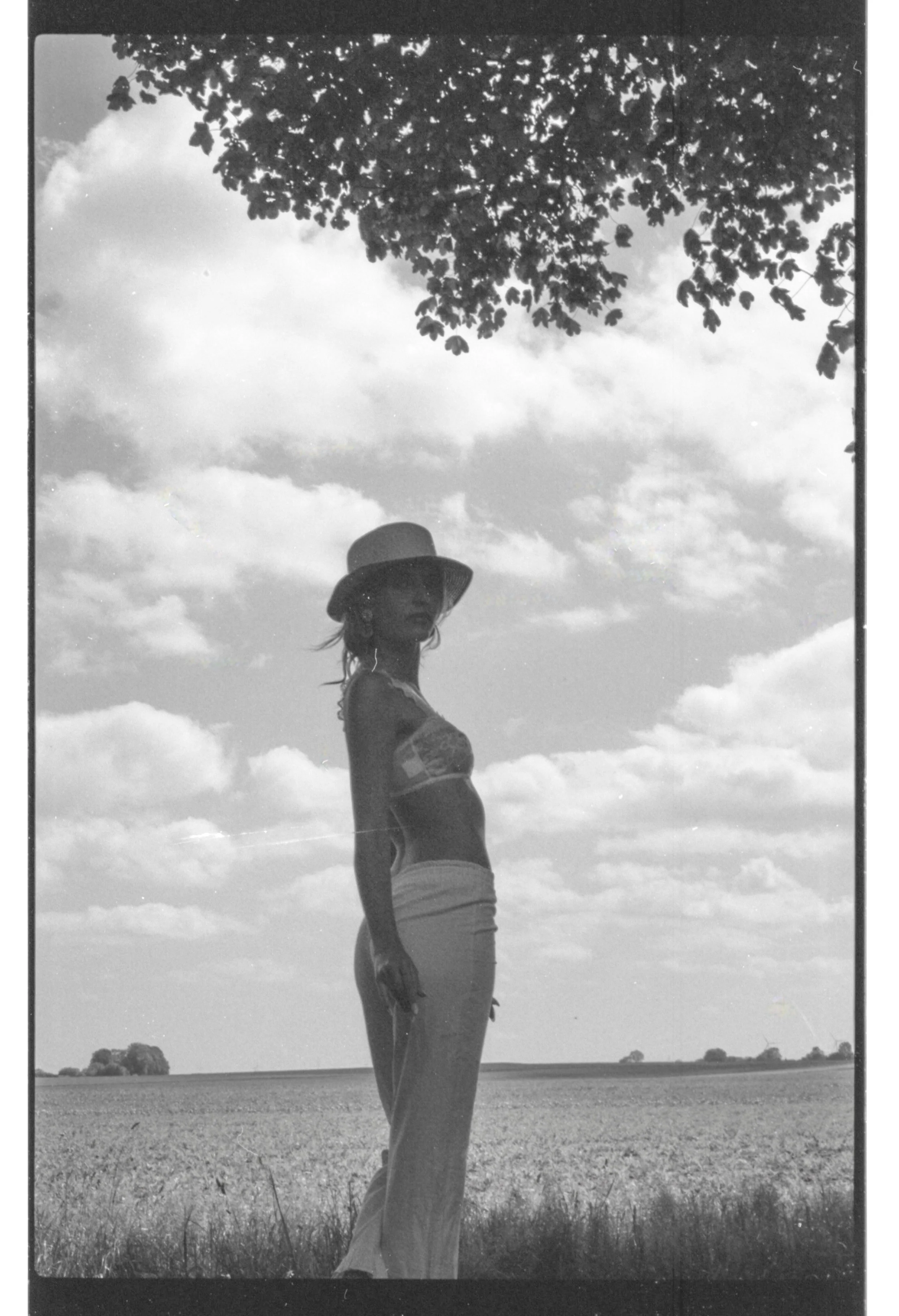 Une femme en kimono, portant un chapeau, se tient dans un champ avec le ciel et des nuages en arrière-plan, vue en noir et blanc.