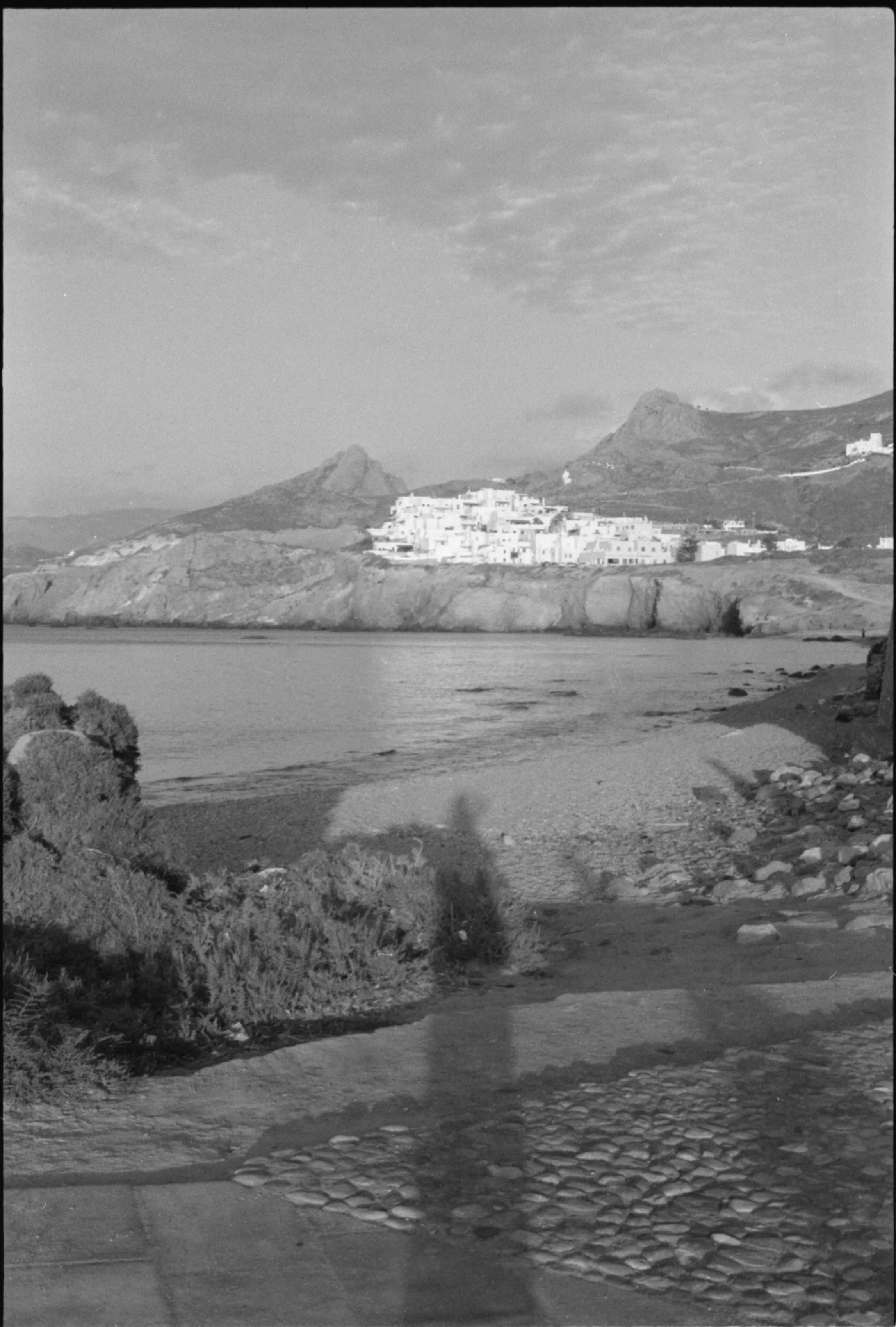 Photo en noir et blanc d'une plage avec des rochers et de l'eau, avec un village blanc en altitude entouré de montagnes en fond.