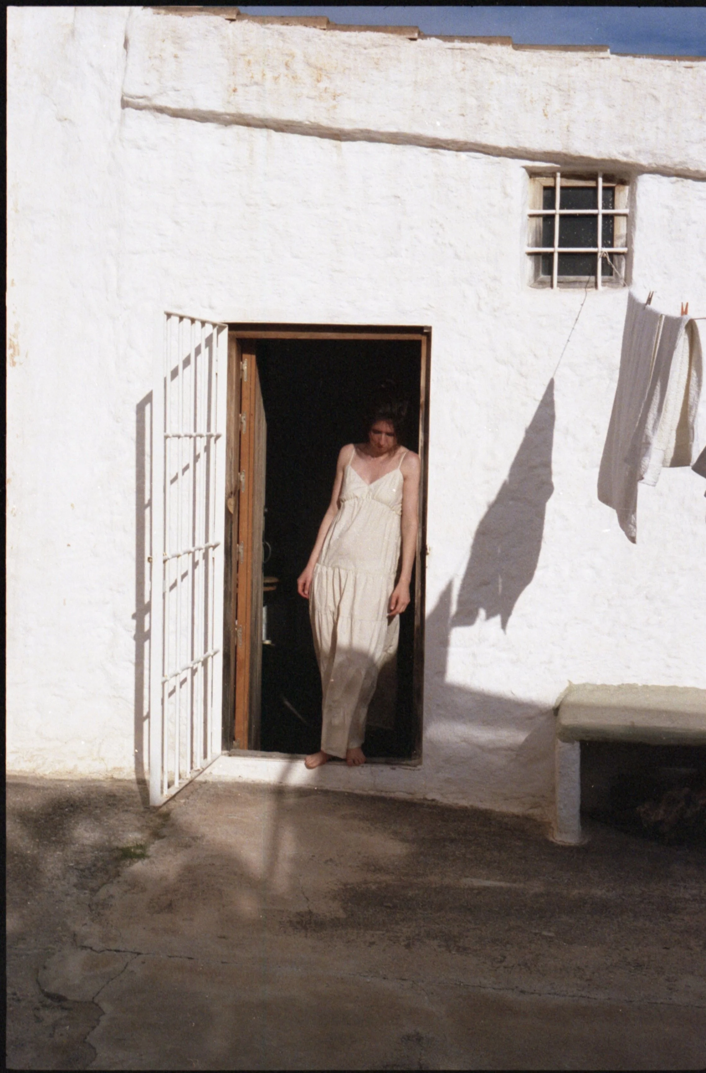 Femme en robe légère debout dans une porte, soleil brillant, effet de lumière et ombres sur le mur blanc