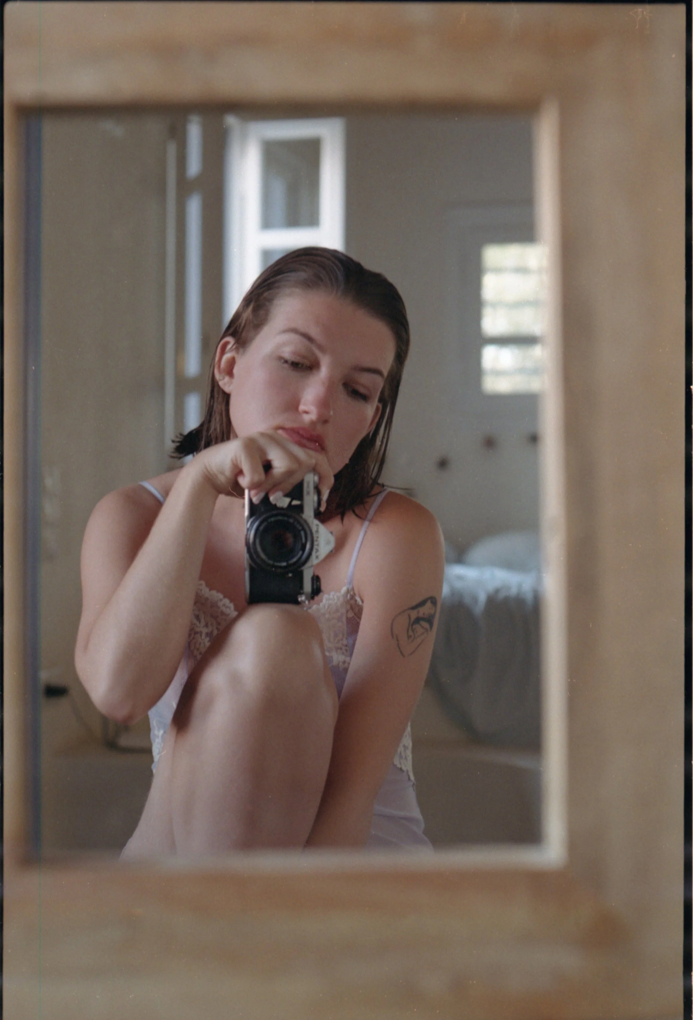 Une femme prend une photo d'elle-même dans un miroir. Elle a les cheveux humides, porte une camisole en dentelle blanche, et a un tatouage d'une tête de lion sur le bras droit.