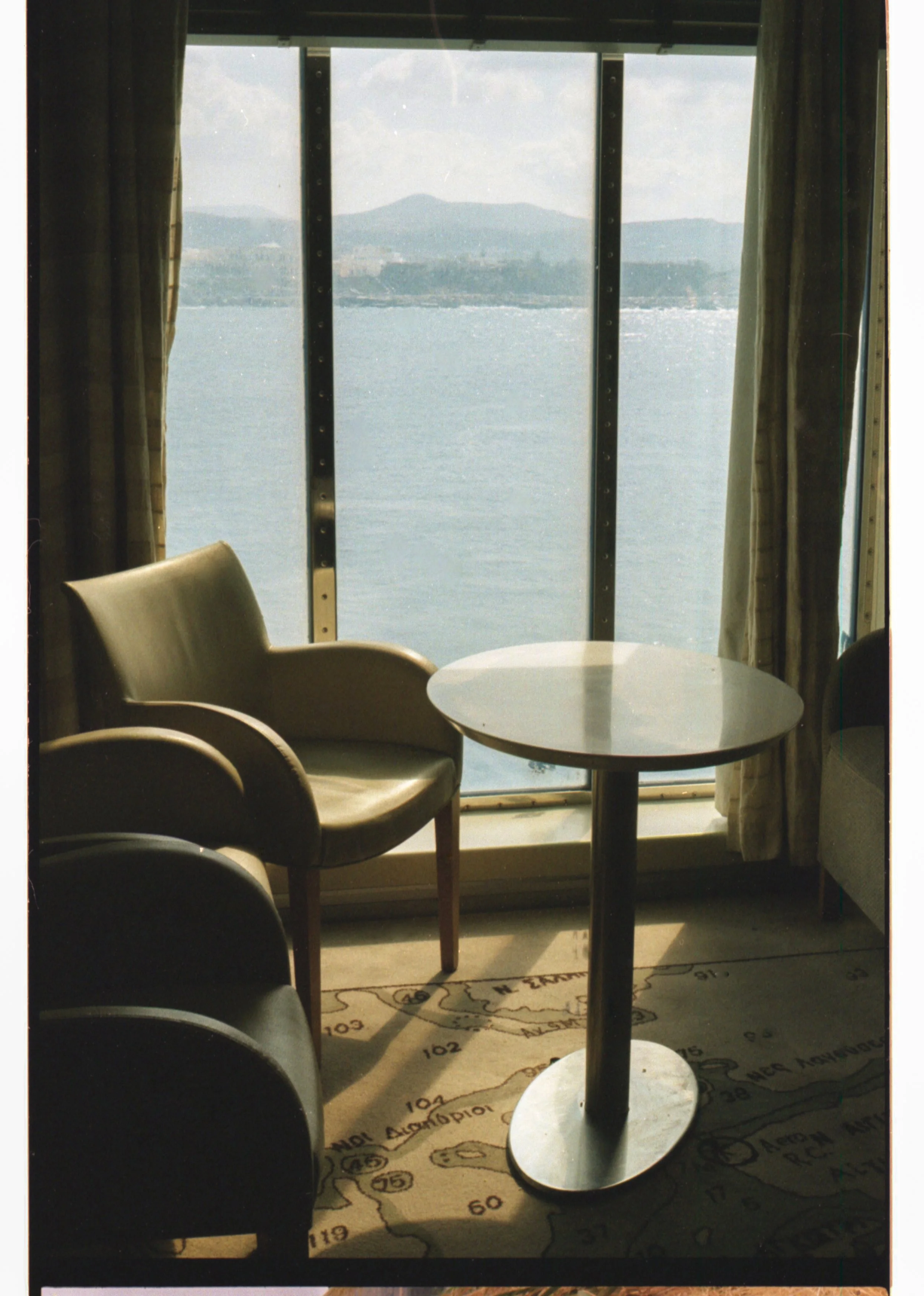 Une vue depuis une salle avec des fauteuils et une table, offrant une vue sur un plan d'eau et des montagnes à travers de grandes fenêtres.