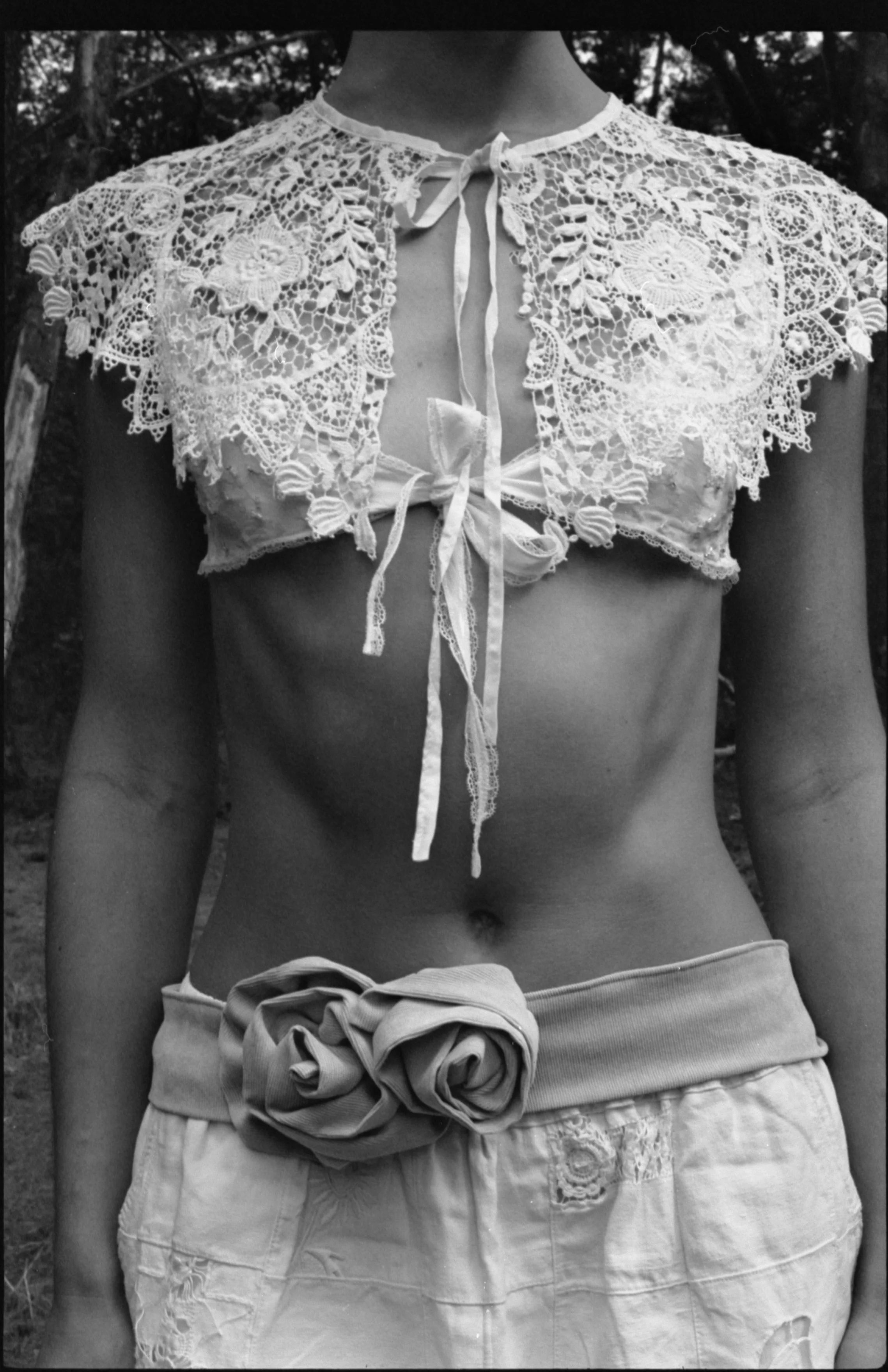 Une femme porte un haut en dentelle et un pantalon avec une grosse fleur en tissu à la taille, image en noir et blanc.
