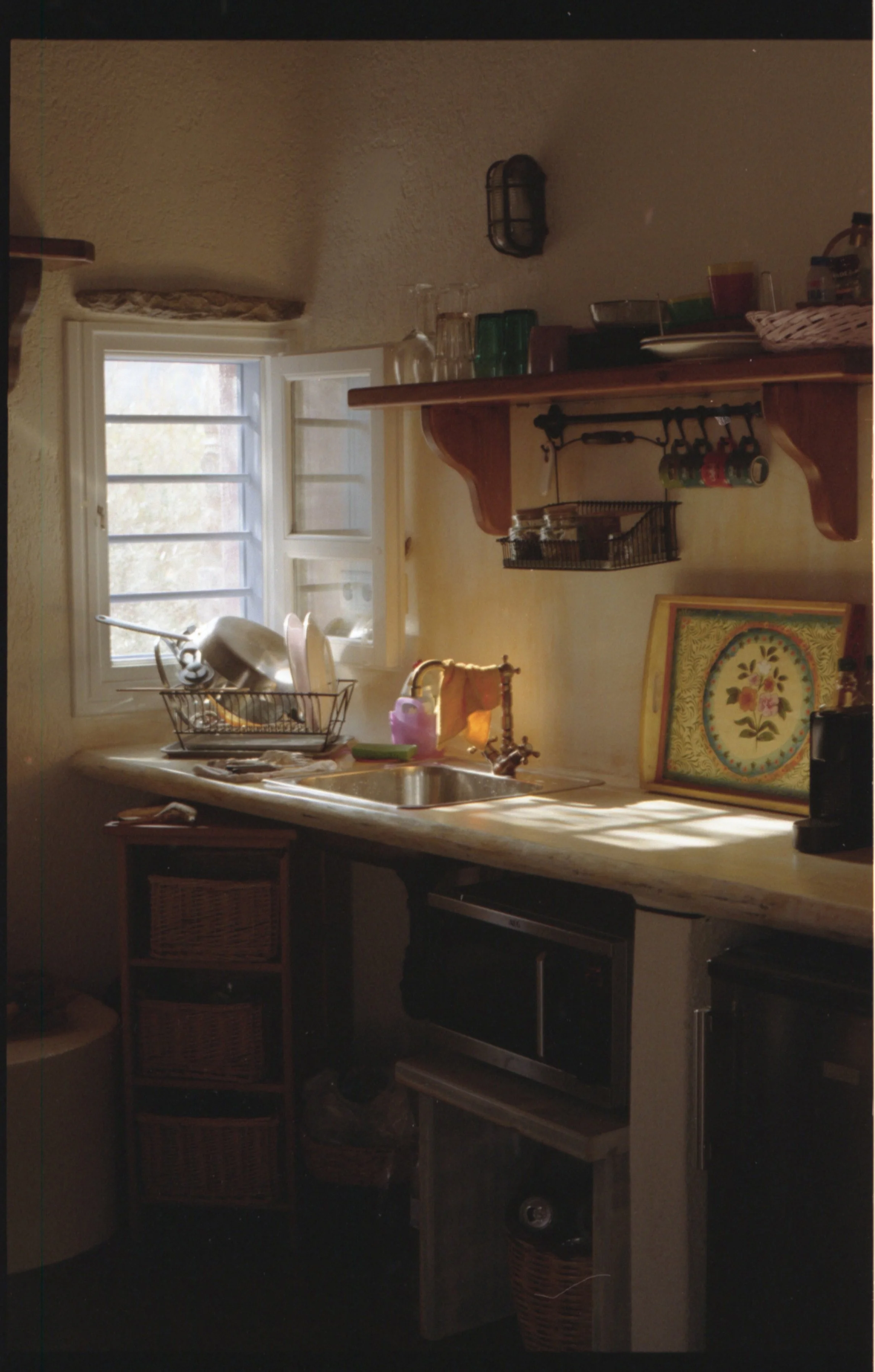 Une cuisine rustique avec une fenêtre donnant sur l'extérieur, un évier, des étagères en bois avec des bocaux et des assiettes, un plateau décoratif avec un motif floral, et des ustensiles de cuisine.