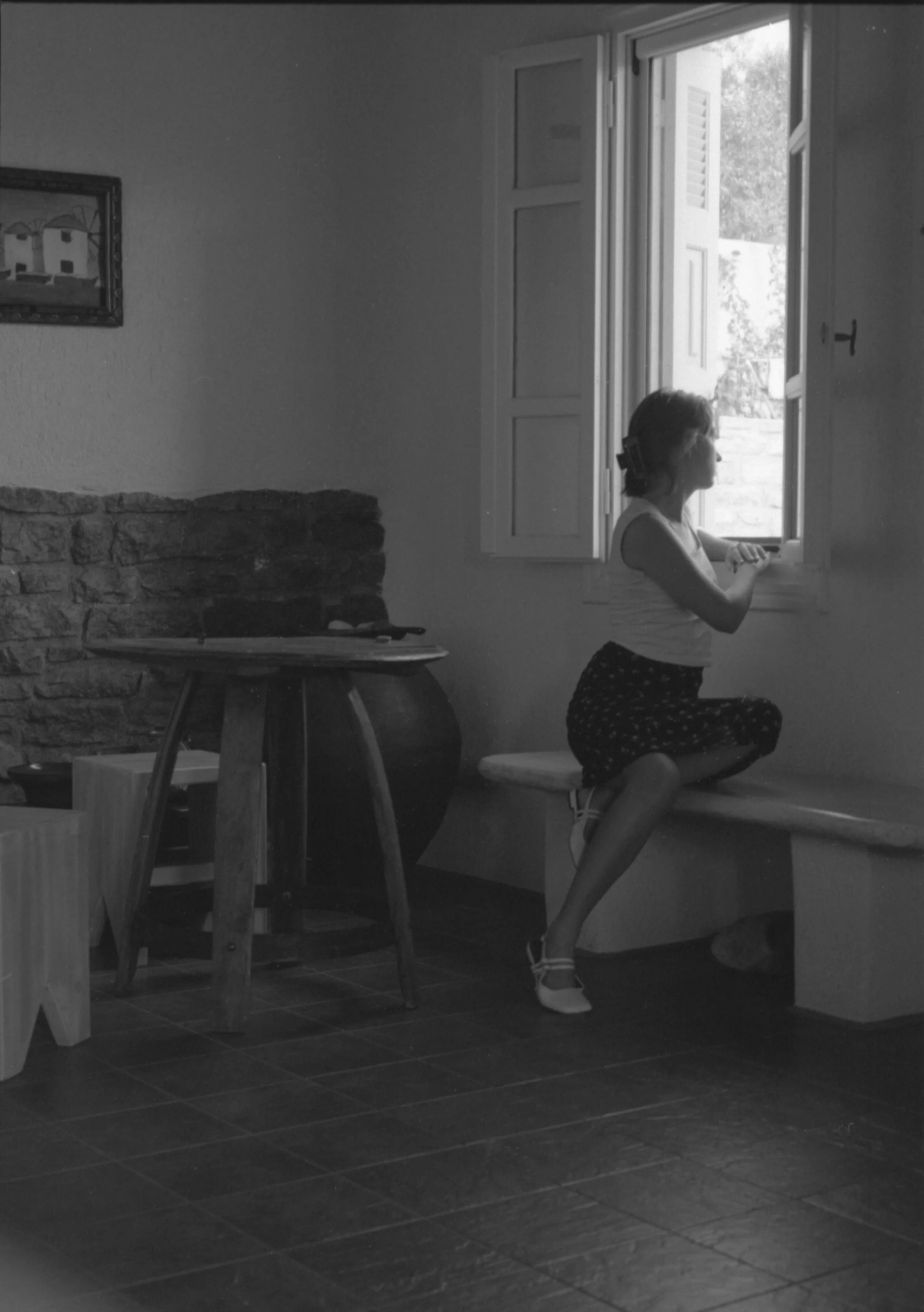 Une jeune femme assise sur un banc en plomb observant par une fenêtre ouverte dans une pièce. La pièce a un sol en carreaux foncés, une table en bois, et un mur en pierre.