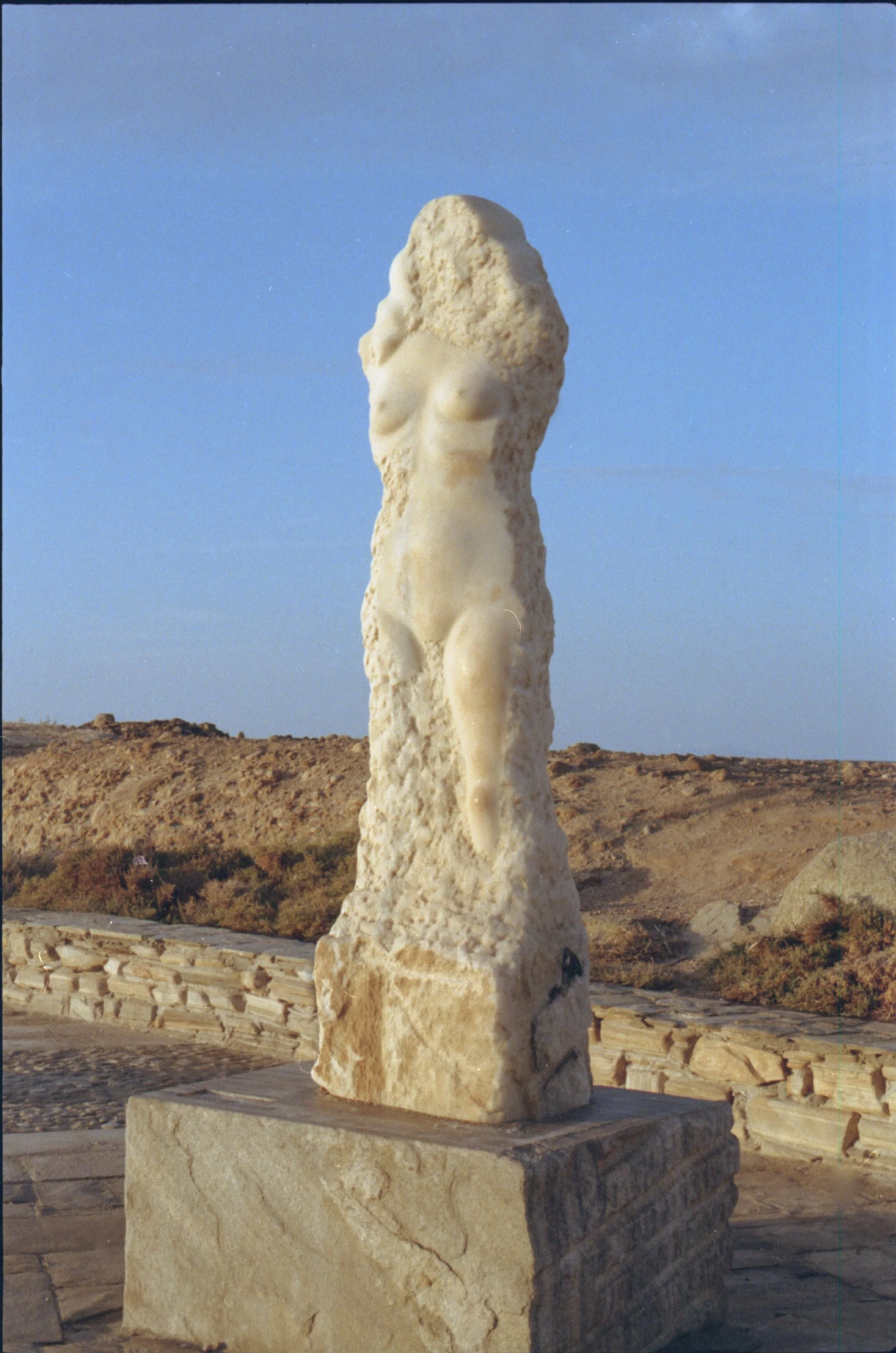 Statue en pierre représentant un corps féminin partiellement effacé, avec un fond de paysage désertique et un ciel bleu.