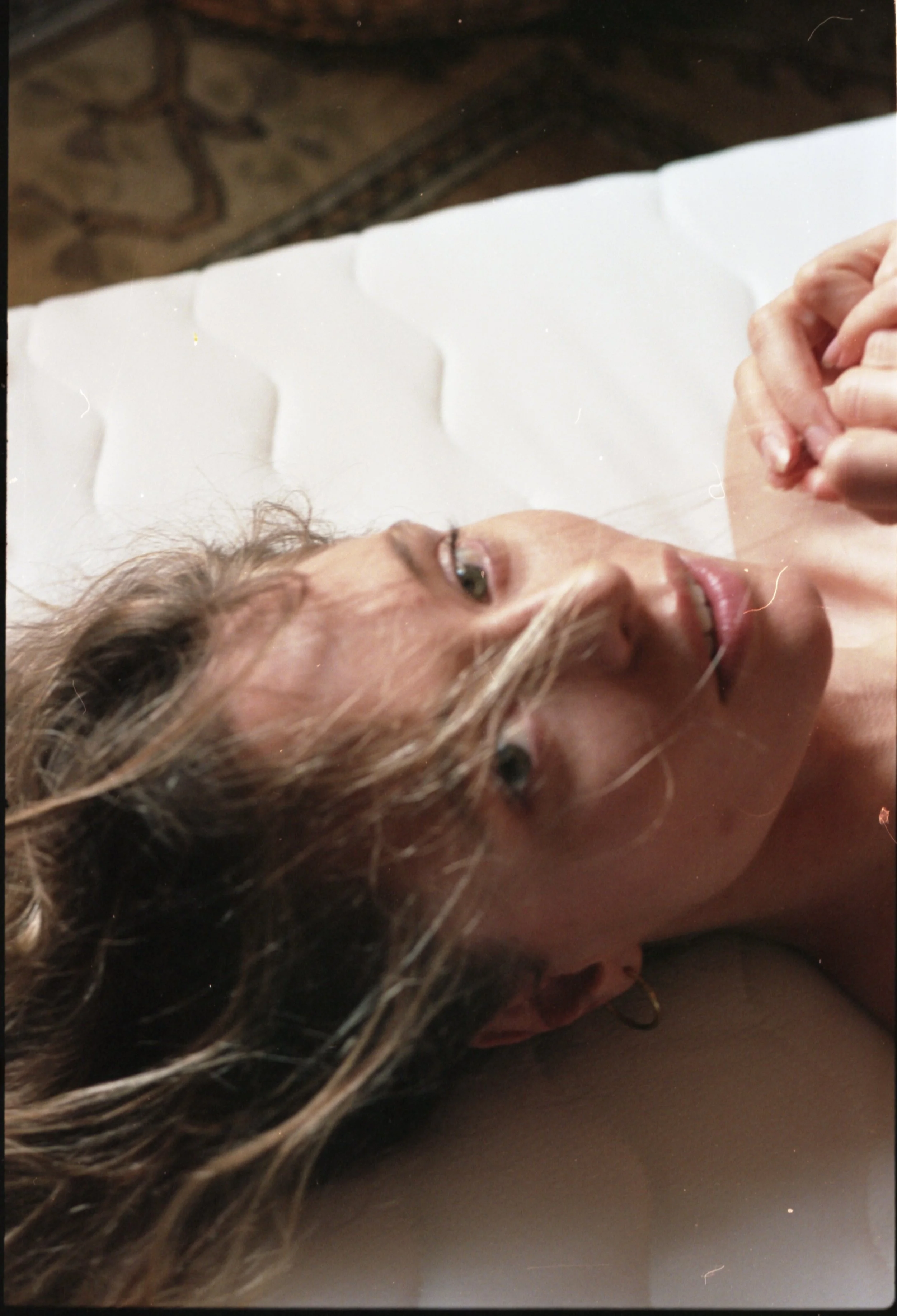 Une femme allongée sur un lit avec des cheveux longs et ondulés, regardant le haut, avec un léger sourire. Elle tient ses mains ensemble près de sa poitrine.