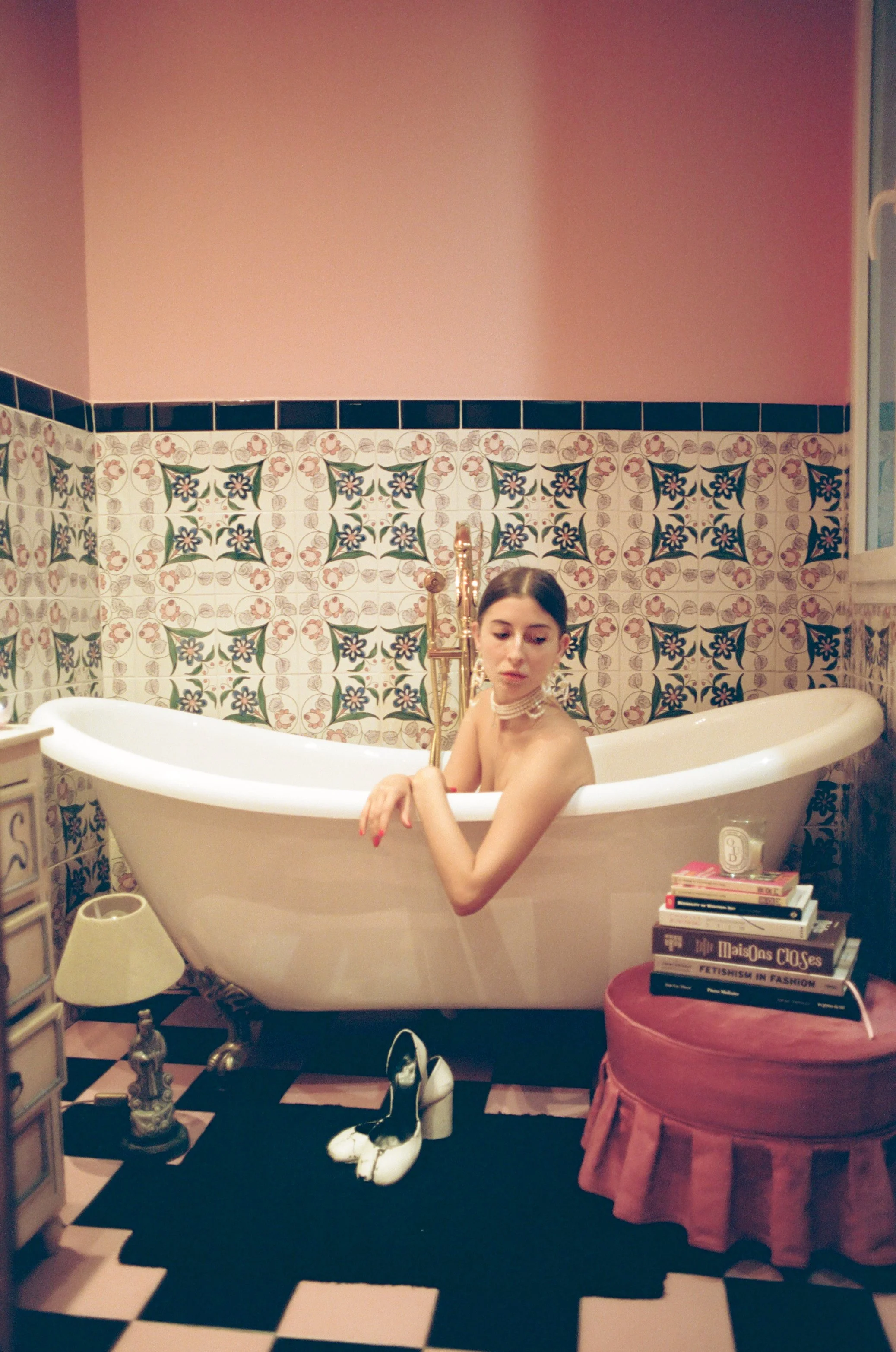 Une jeune femme dans une baignoire, entourée d'éléments décoratifs et de livres, dans une pièce au style vintage, avec un plancher à carreaux noirs et blancs.