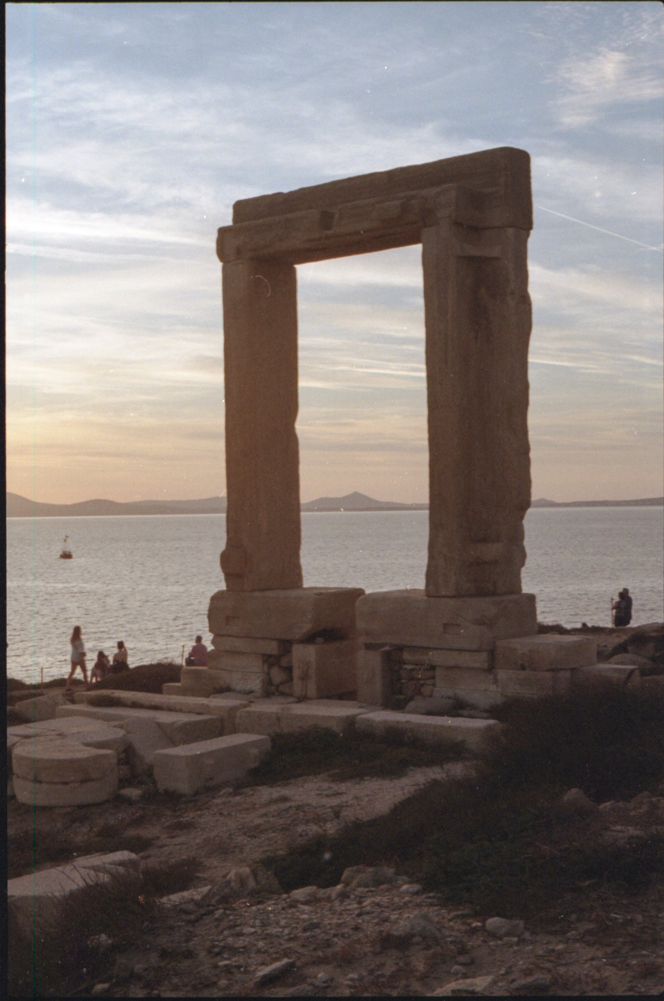 Ruines d'un ancien site archéologique avec un cadre en pierre, au bord d'une mer au coucher du soleil, avec des personnes en arrière-plan.