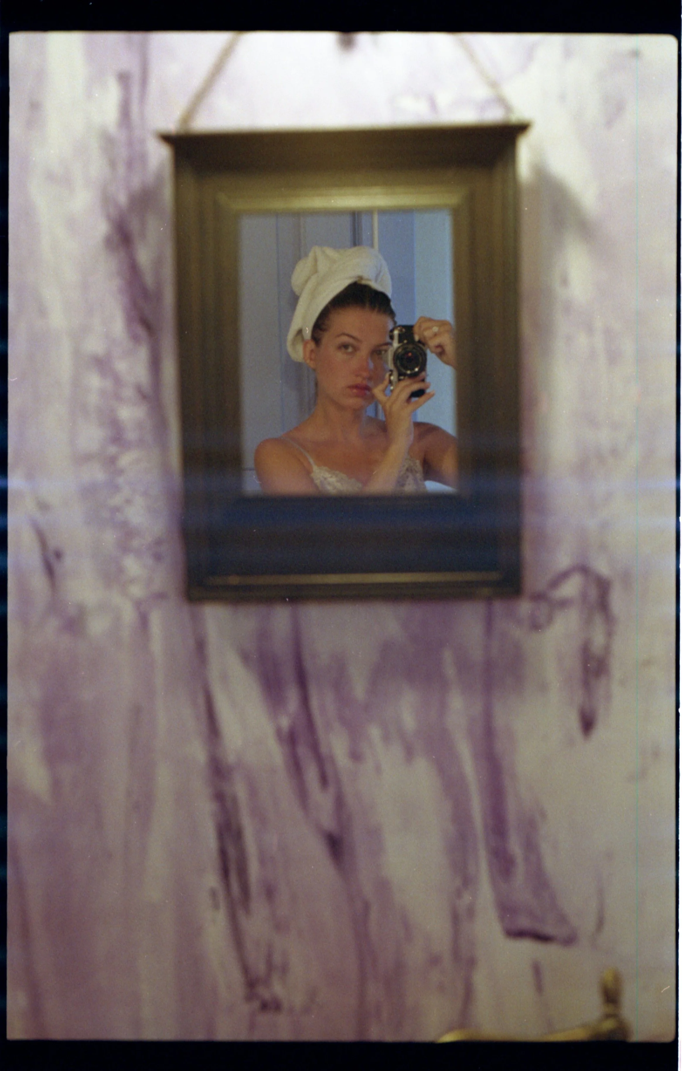 Une femme se regarde dans un miroir, portant une serviette blanche sur la tête et un vêtement léger. Elle tient un appareil photo devant son visage.