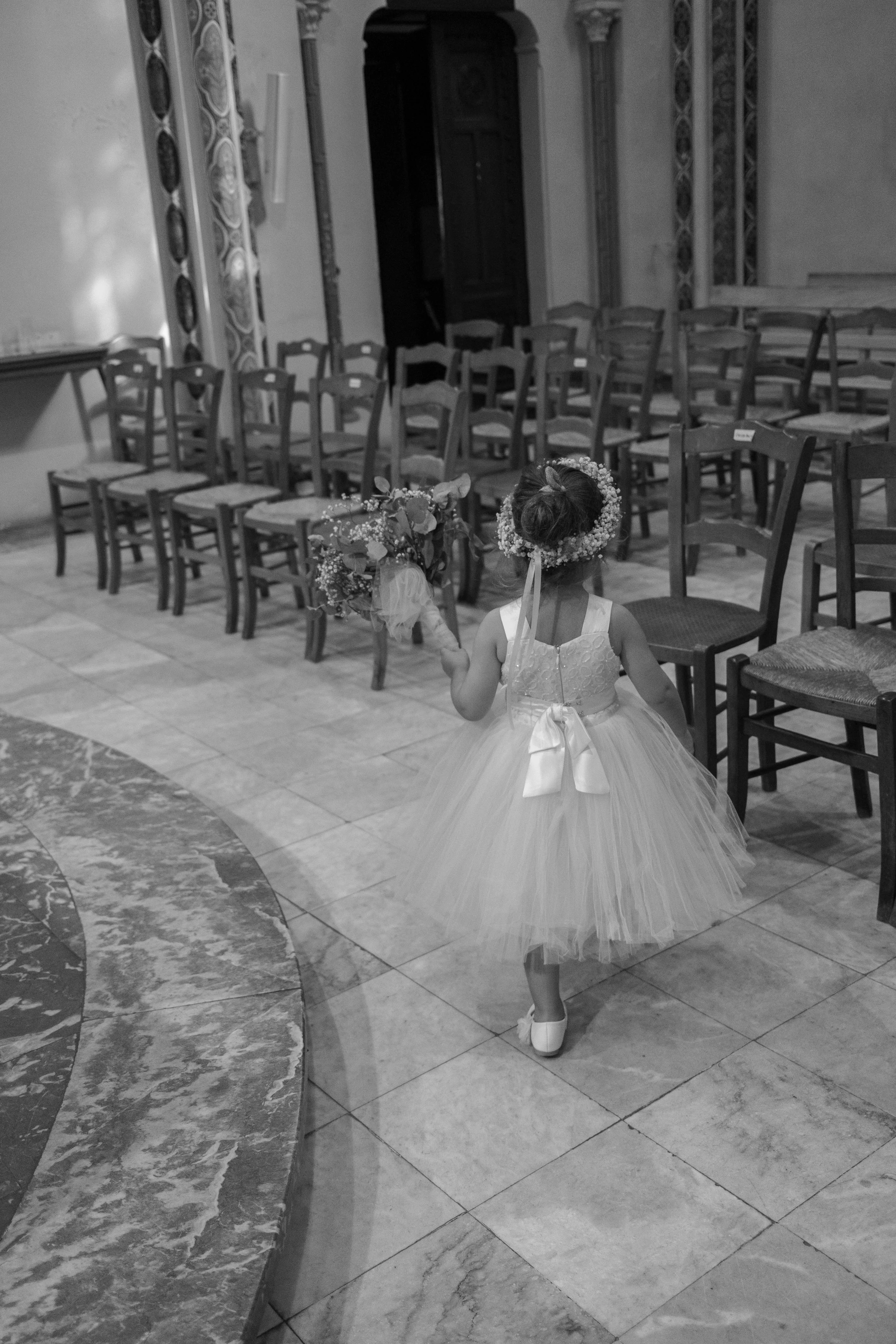 Une petite fille en robe de mariée avec une couronne de fleurs, tenant un bouquet de fleurs, marche dans une salle ornée de chaises en bois.