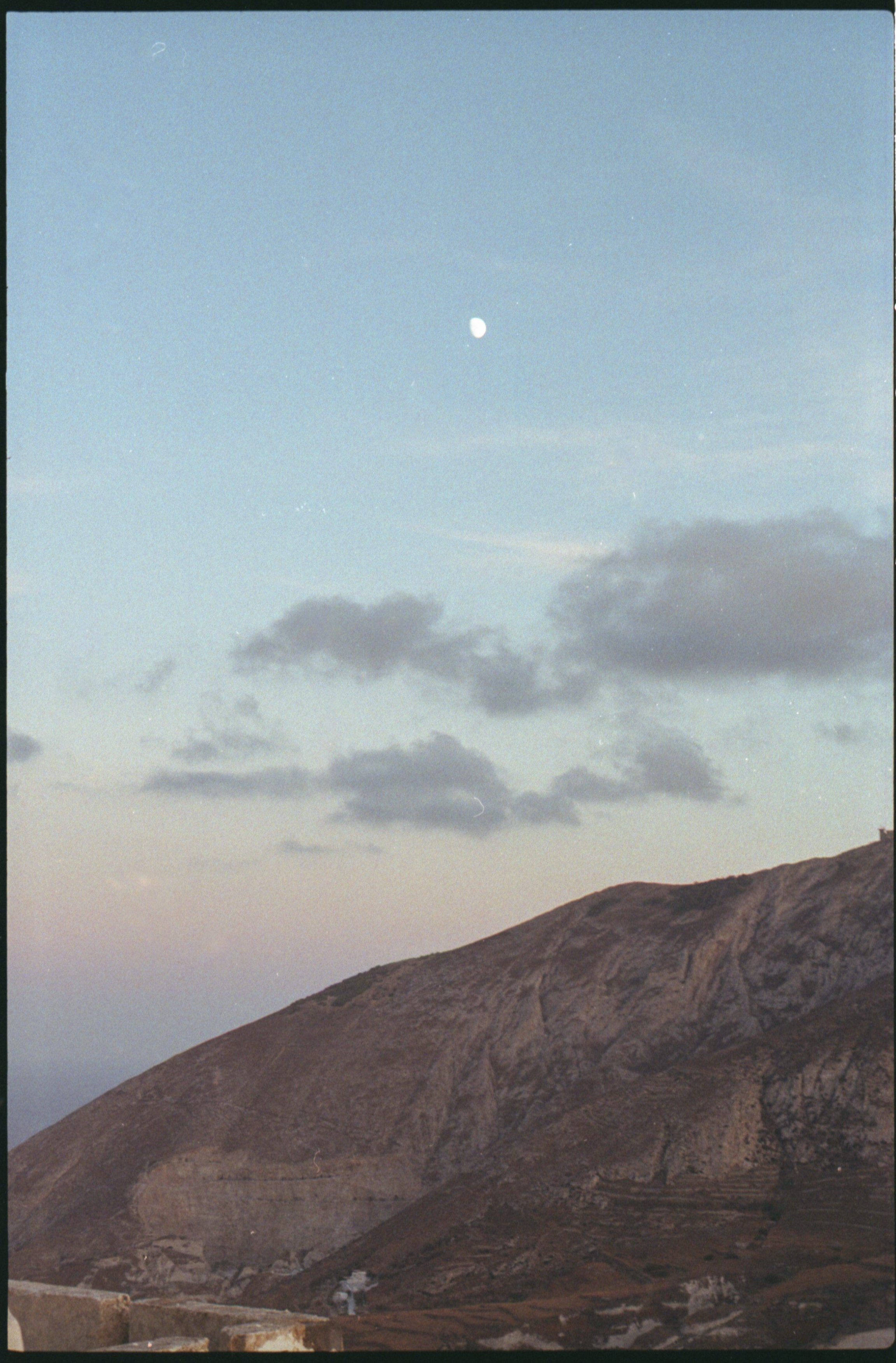 Un paysage de montagne avec un ciel bleu clair, quelques nuages, et la lune visible dans le ciel au crépuscule.