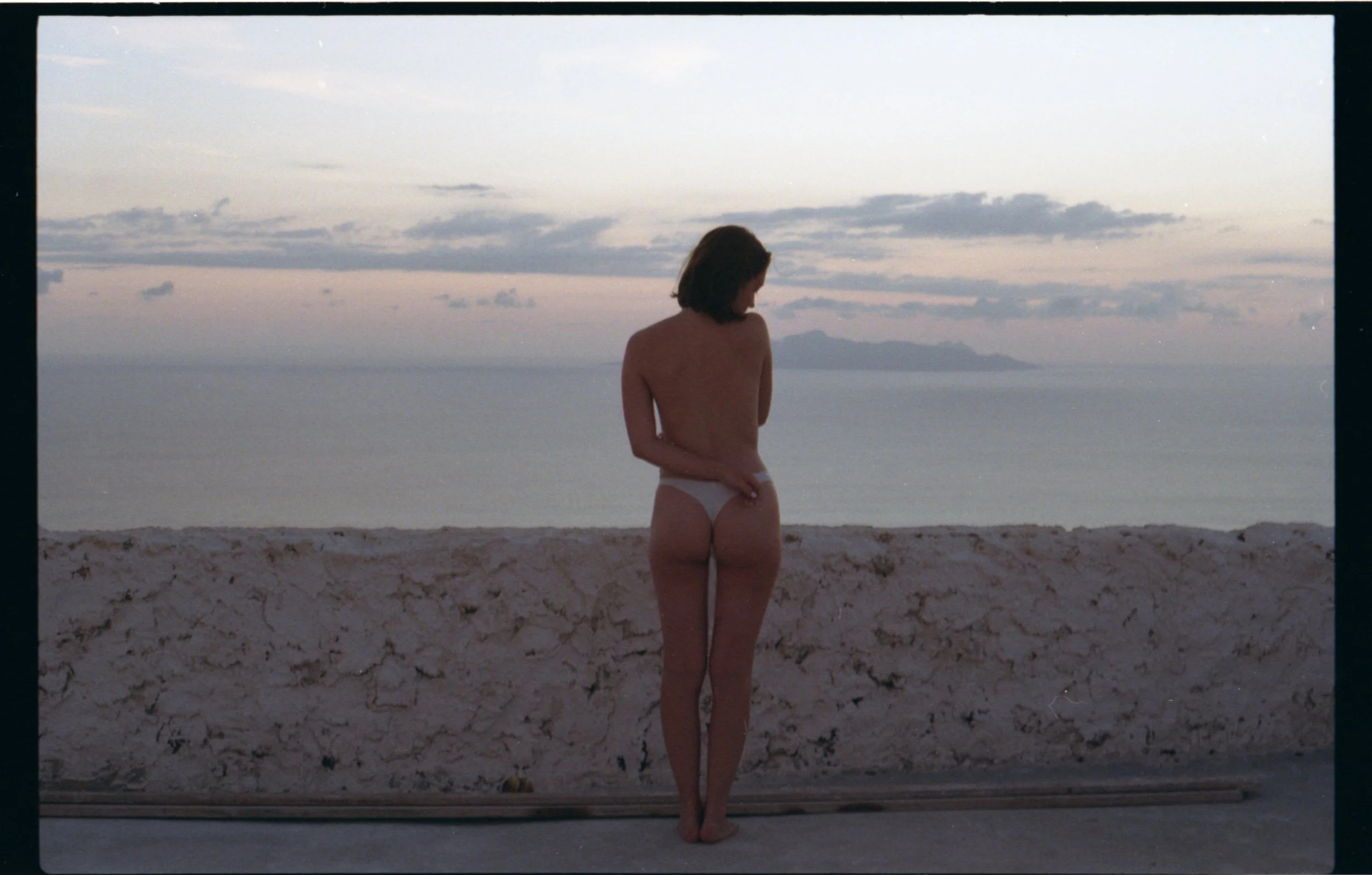 Une femme nue vue de dos, portant uniquement un sous-vêtement blanc, se tenant sur un trottoir face à la mer au coucher du soleil.