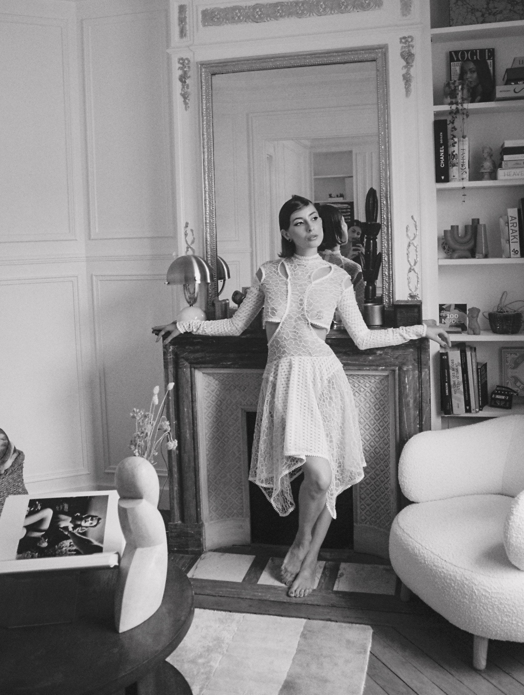 Une femme posant près d'un miroir dans un intérieur élégant, en noir et blanc, portant une robe en dentelle.