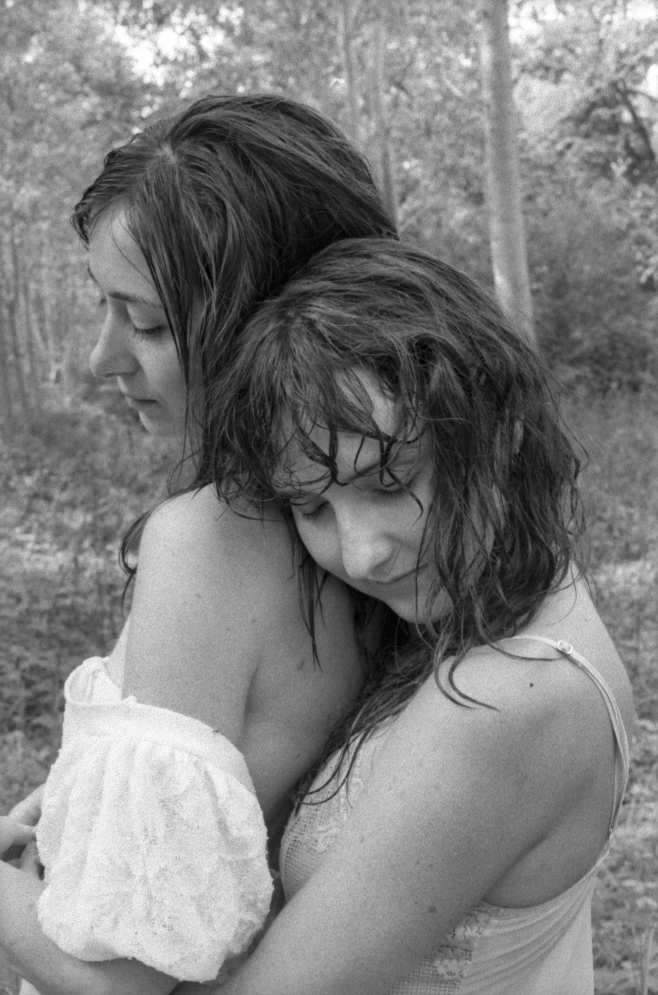 Deux femmes, une avec les cheveux longs et mouillés, l'autre avec les cheveux courts, s'enlacent tendrement dans un environnement naturel forestier.