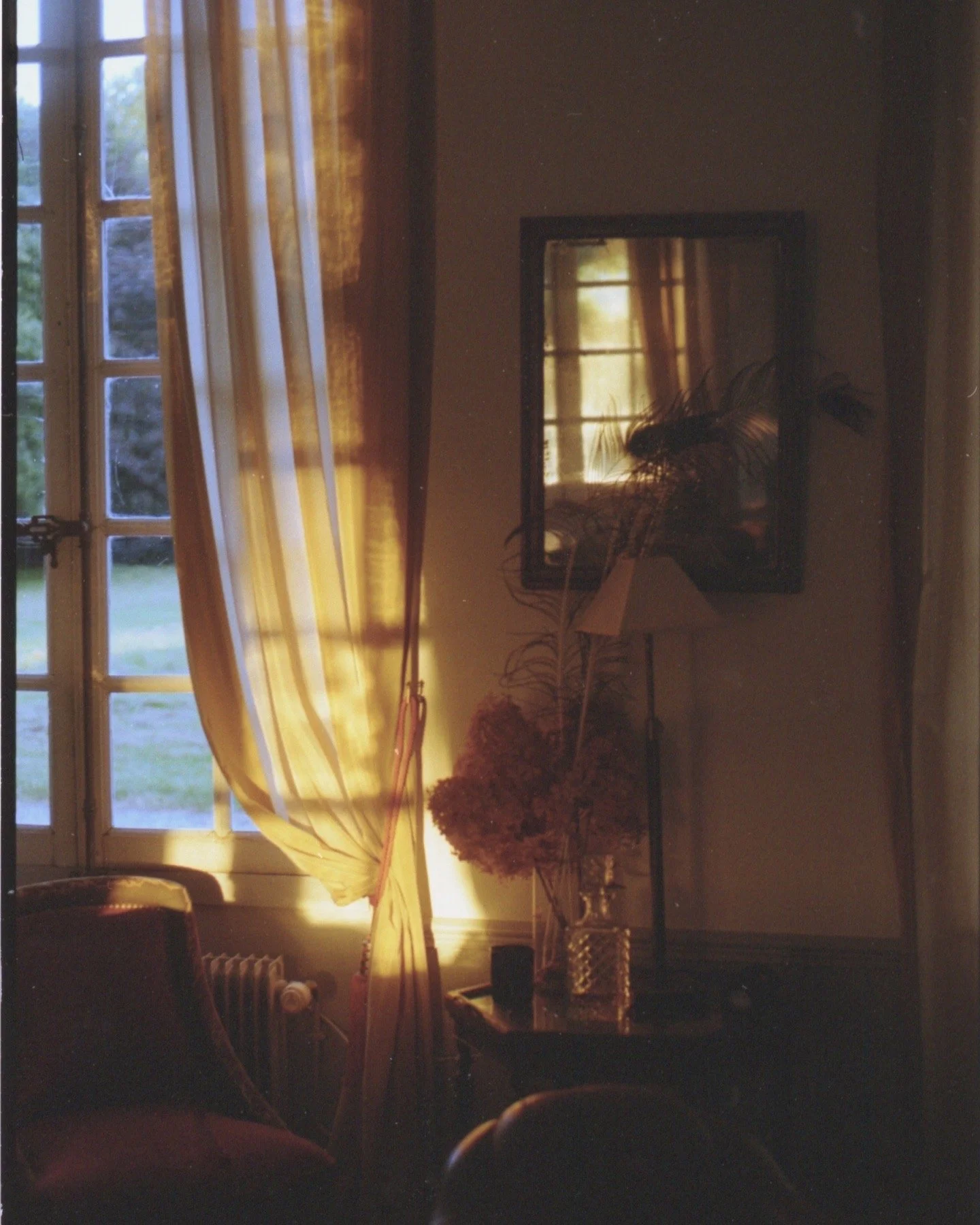 Intérieur d'une pièce avec une grande fenêtre laissant passer la lumière du soleil, des rideaux jaunes, un miroir au mur, un lampadaire, une table avec un vase contenant des fleurs séchées ou une plante, et un fauteuil.