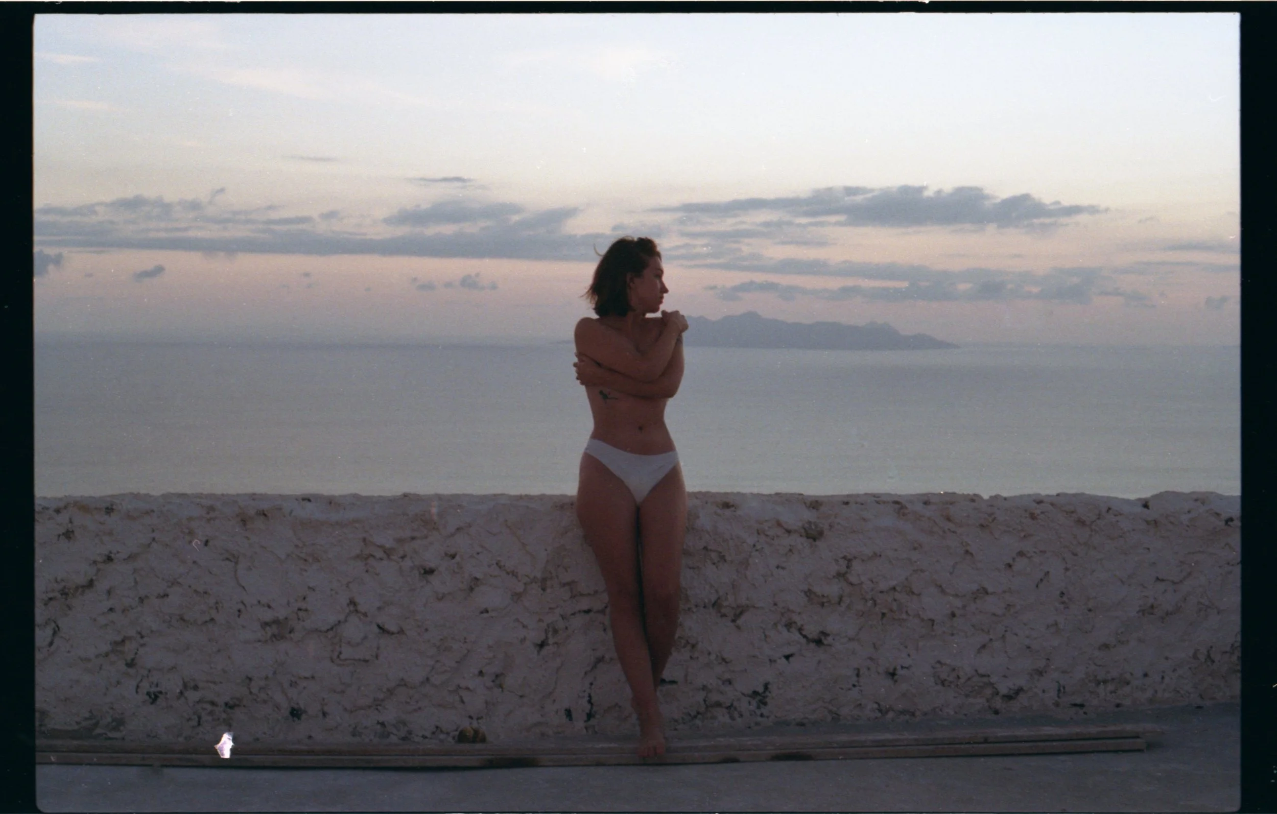 Femme en sous-vêtements blanc, debout contre un mur en pierre sur une plage au coucher du soleil, regardant vers la mer.