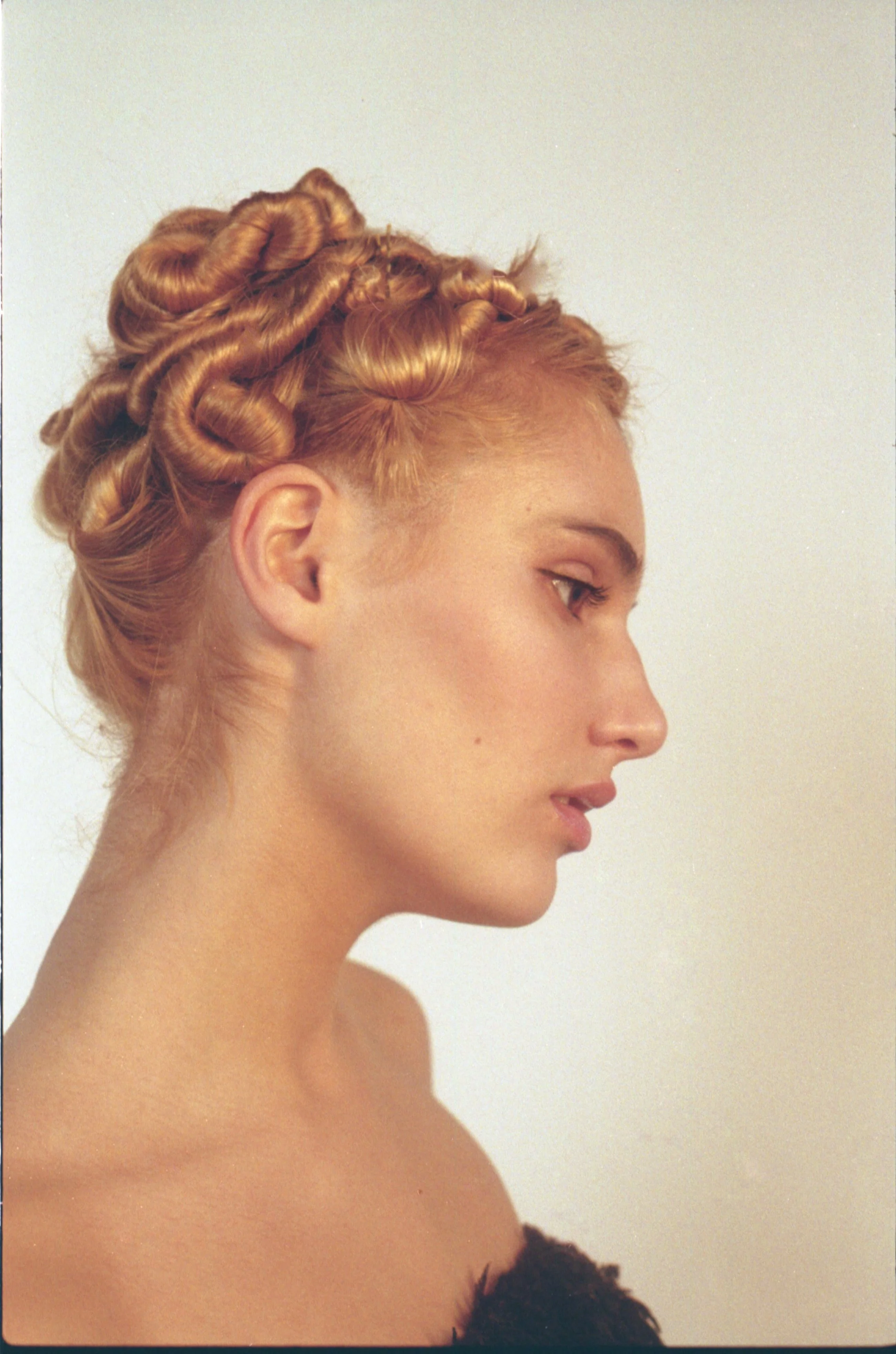 Femme aux cheveux roux coiffés en boucles vintage, vue de profil, avec épaule dénudée contre un fond neutre.