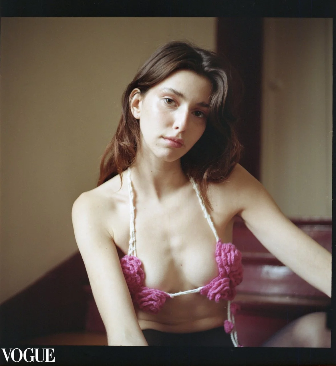 Une jeune femme aux cheveux longs et bruns pose pour une photo, portant un top en crochet rose. Mention VOGUE en bas à gauche.
