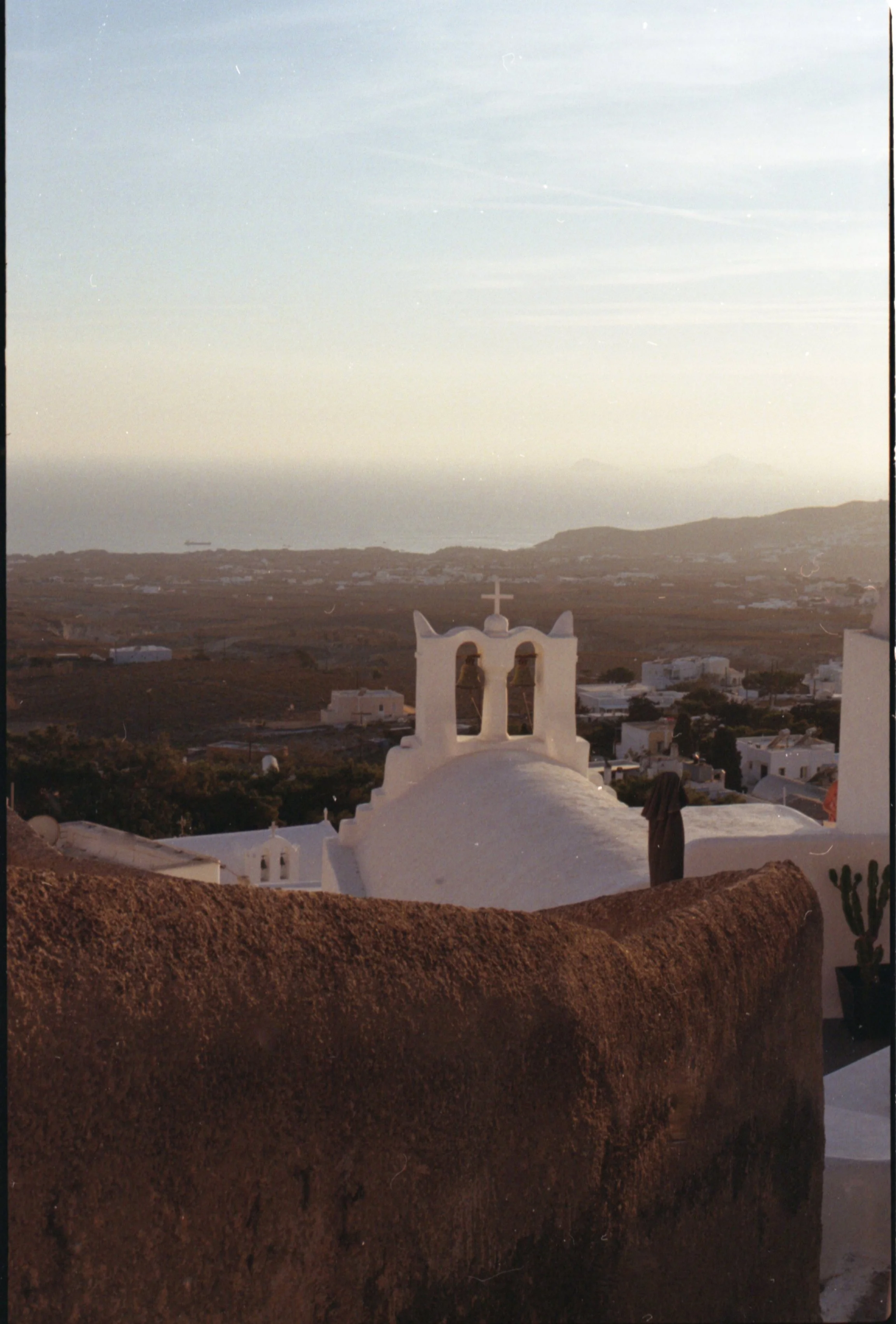 Vue d'une église blanche avec un clocher et une croix, surplombant un paysage rural avec des maisons blanches et les montagnes en arrière-plan, au coucher du soleil.