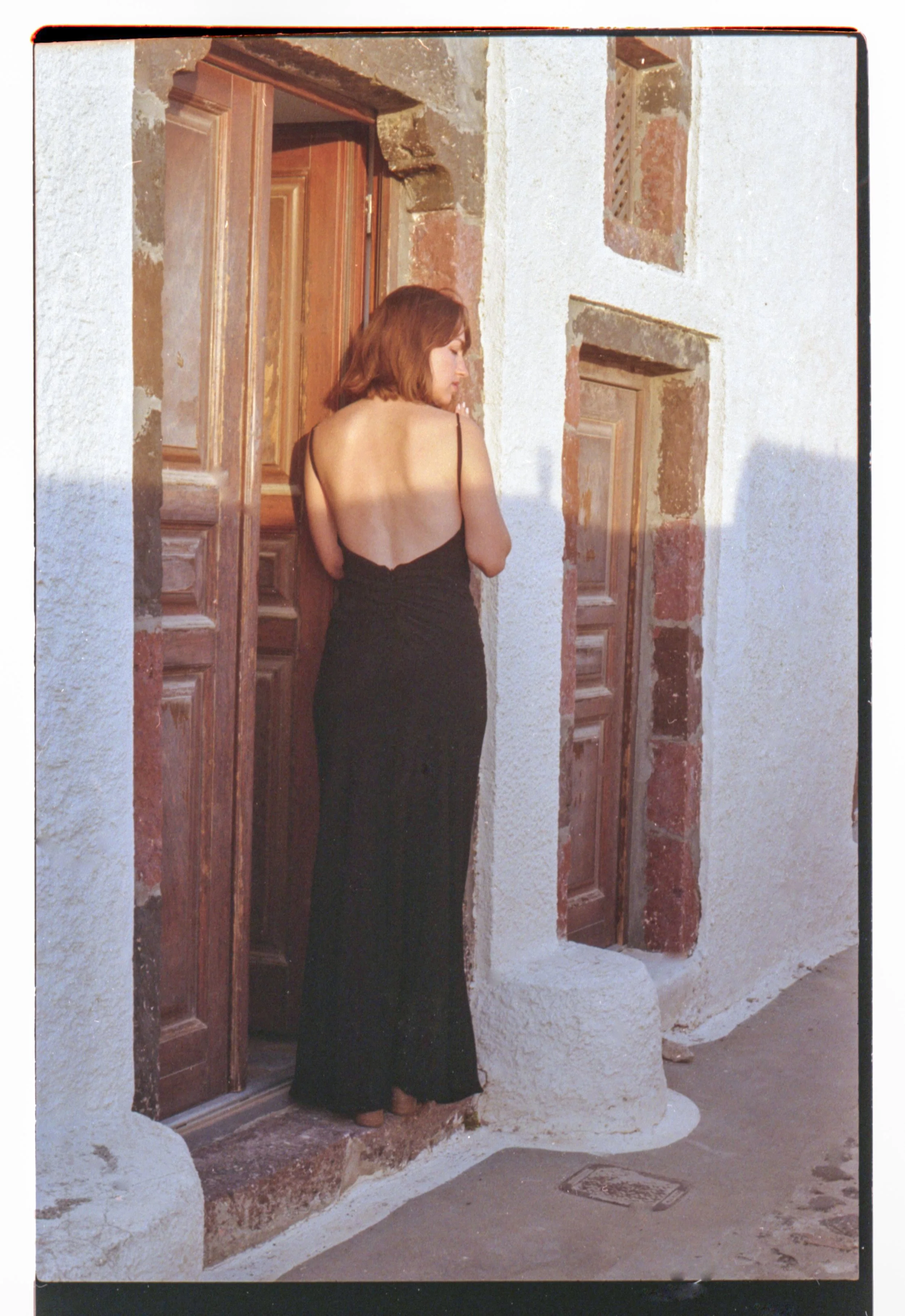 Une femme en robe noire longue et à dos, debout devant une porte en bois sur une façade en pierre. La lumière du soleil crée une ombre sur son dos.