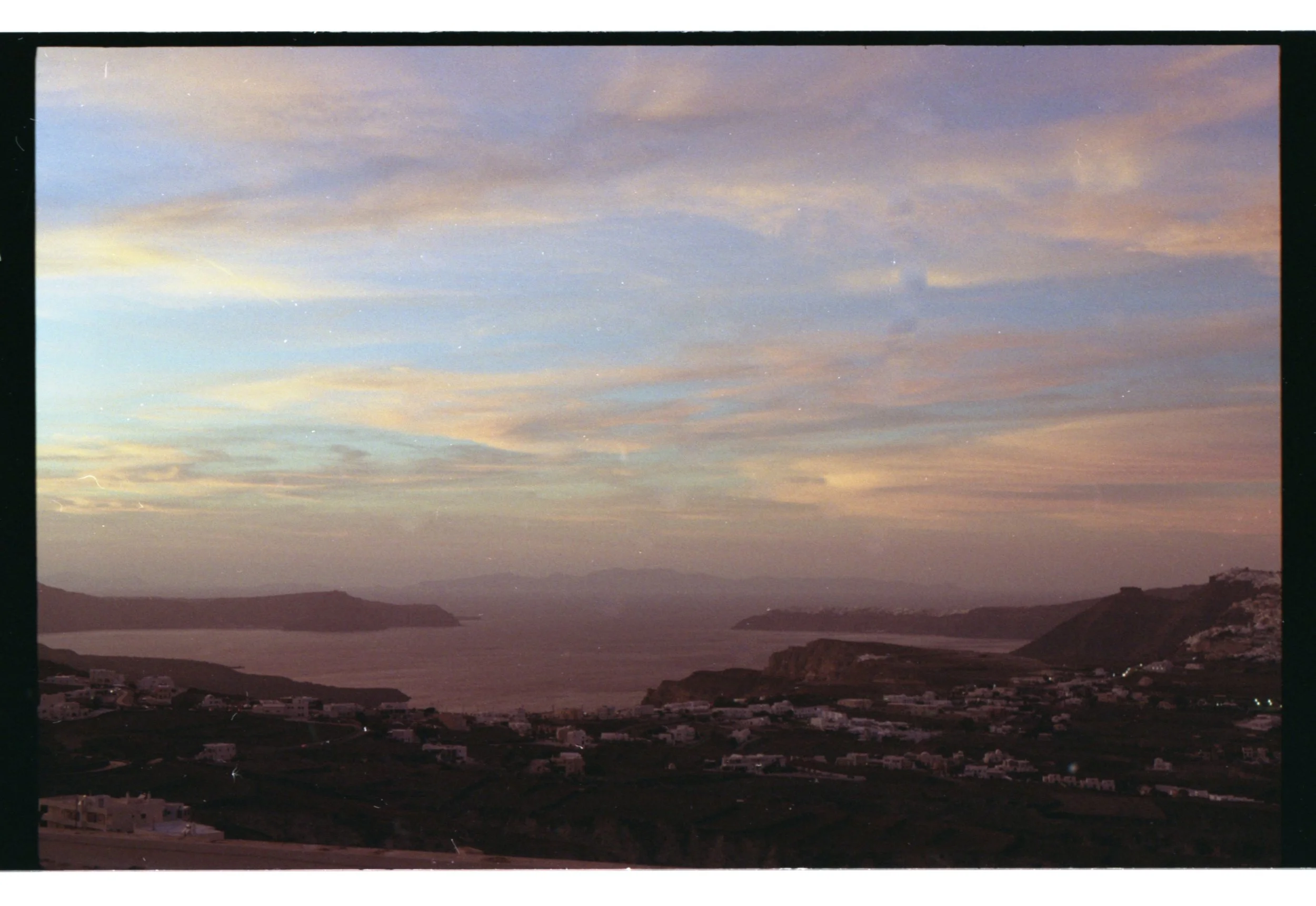 Paysage côtier avec mer, collines et maisons, ciel au crépuscule avec nuages colorés.