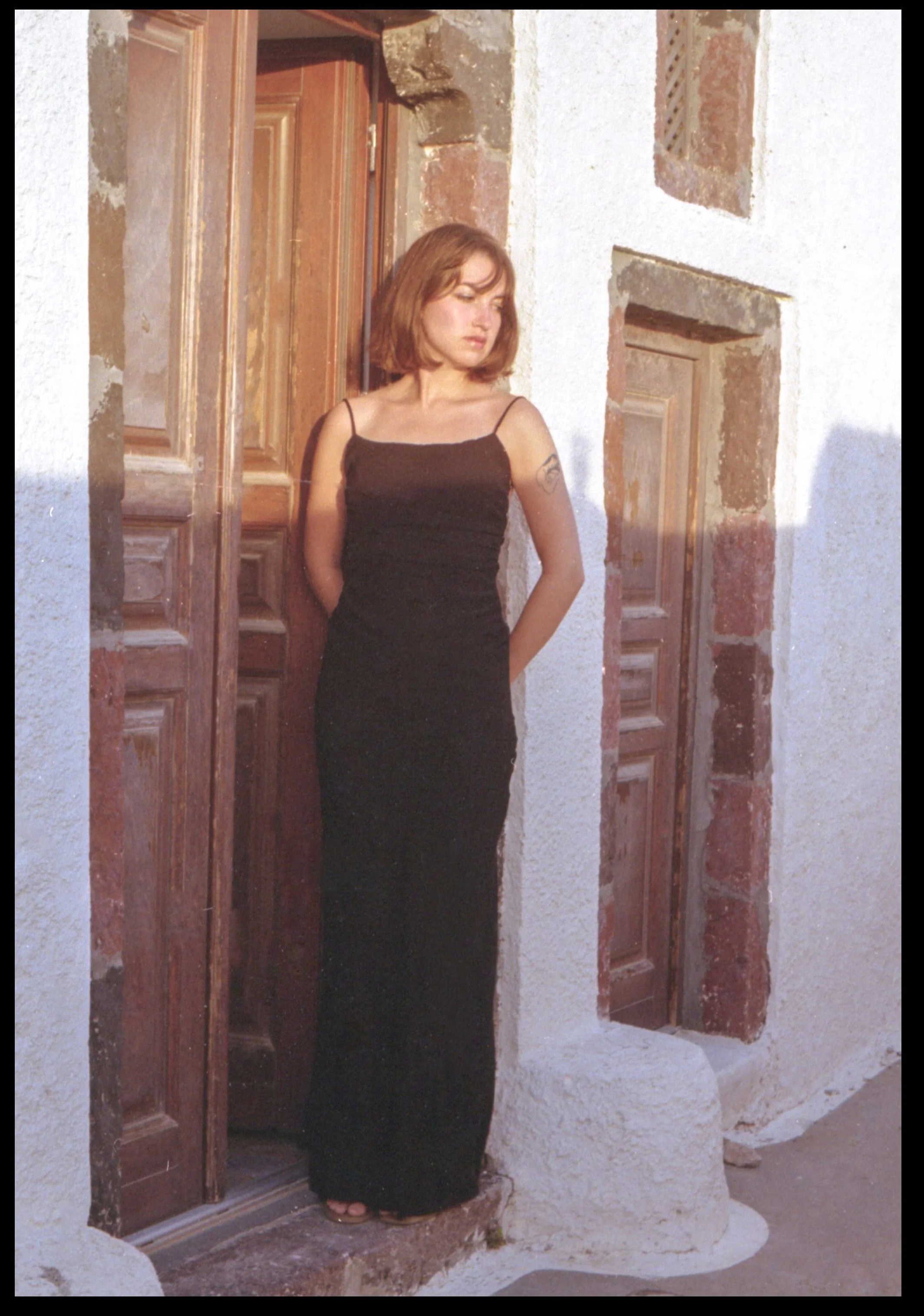 Une jeune femme en robe noire longue, debout dans une porte en bois, laissant la lumière du soleil illuminer son visage et ses épaules, avec un mur blanc en arrière-plan.