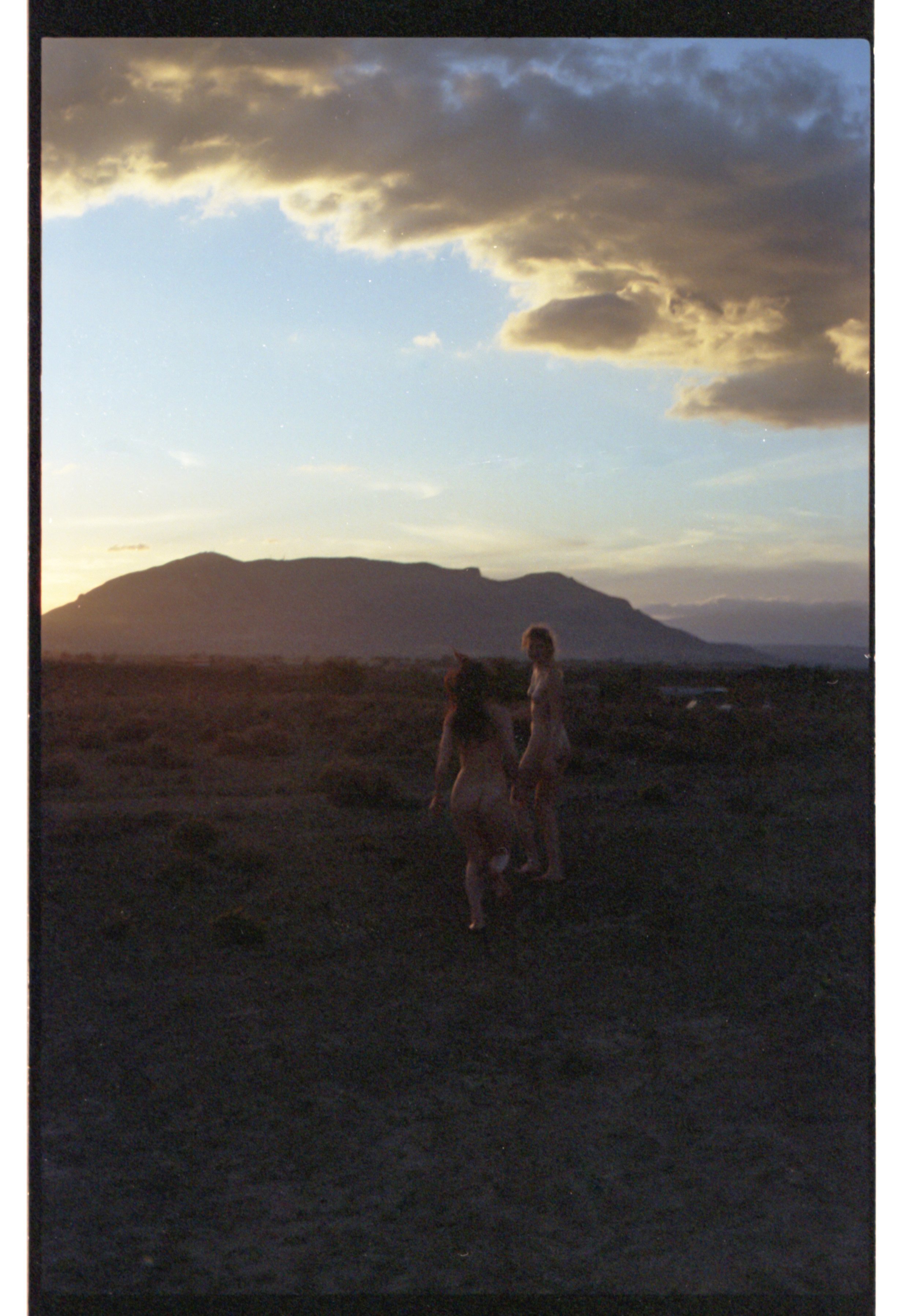 Deux femmes nues se tenant par la main dans un paysage désertique au coucher du soleil, avec des nuages dans le ciel et une montagne en arrière-plan.