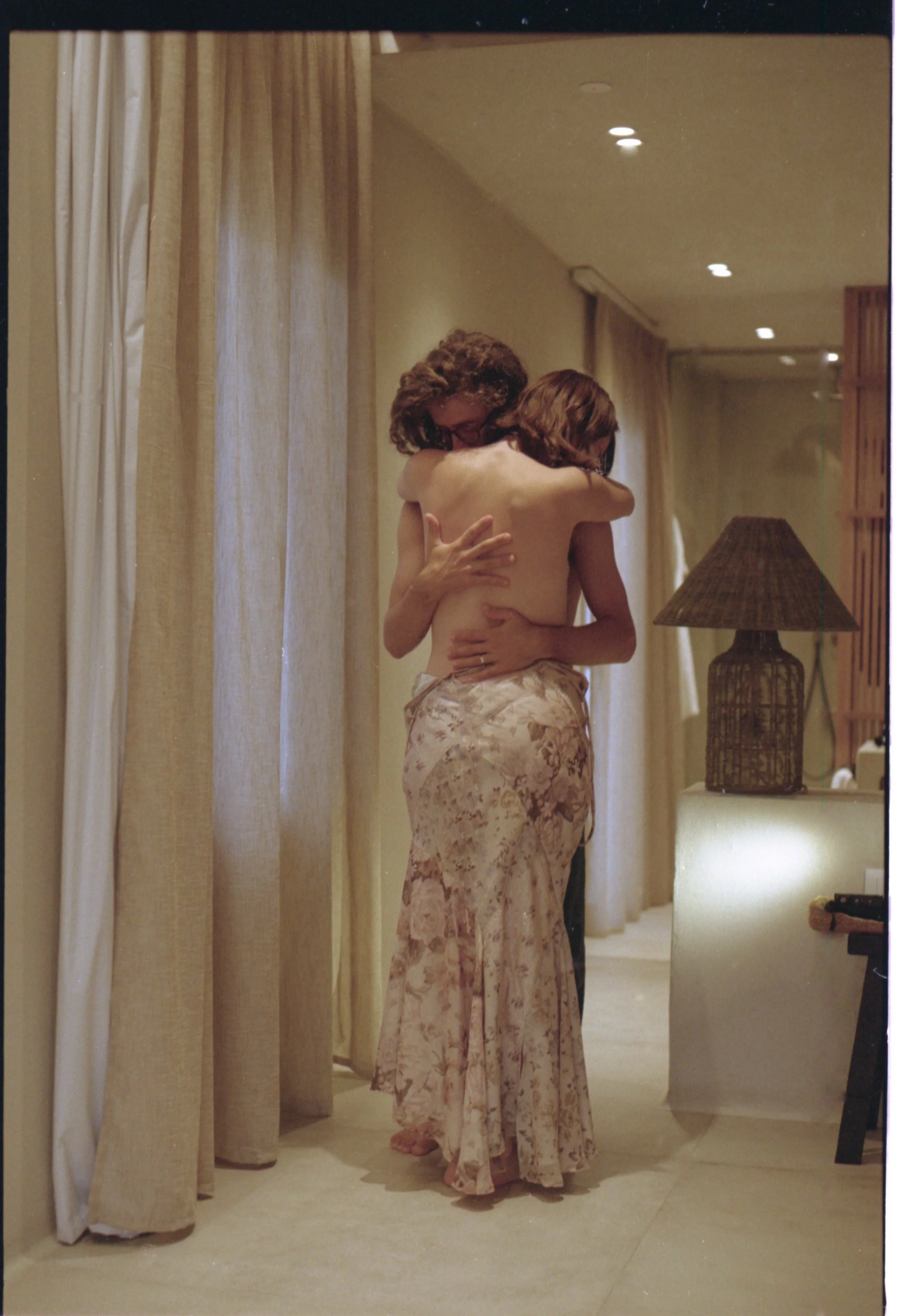 Deux personnes s'embrassent chaleureusement dans une pièce cosy avec des rideaux beiges et une lampe en rotin.