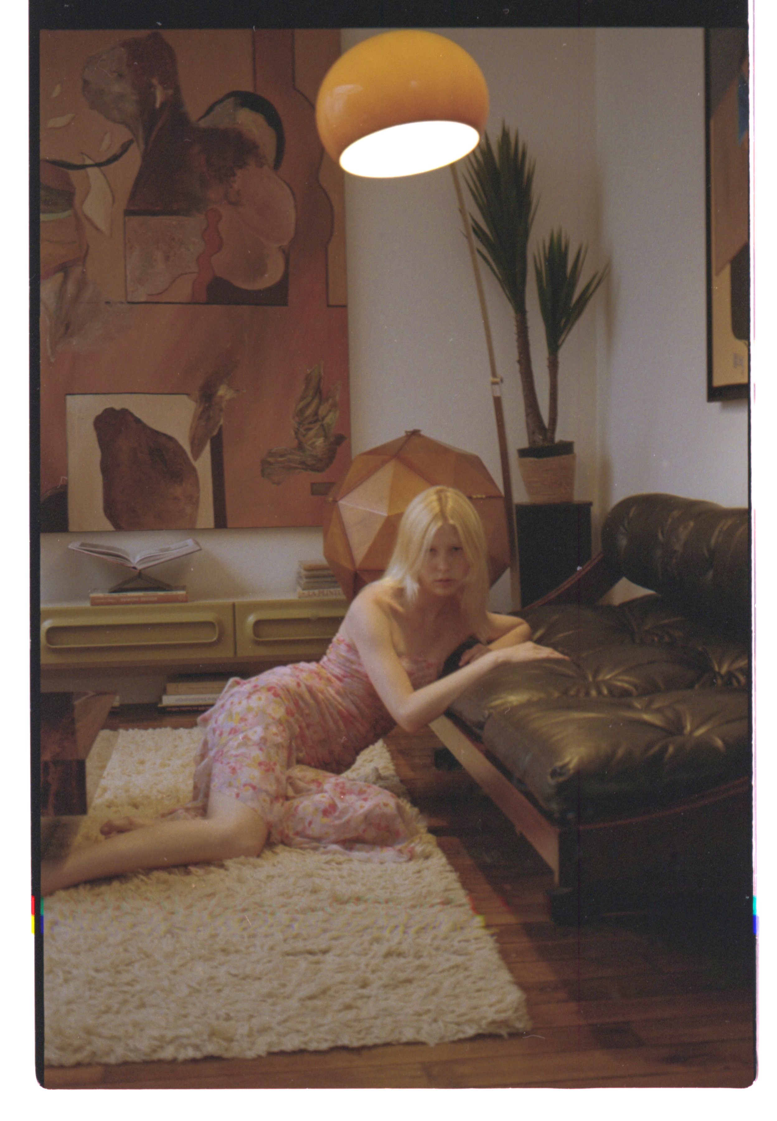 Une jeune femme blonde en robe rose, allongée sur un tapis beige, dans un salon avec un canapé en cuir, une lampe suspendue jaune, une grande plante d'intérieur, et plusieurs œuvres d'art accrochées aux murs.