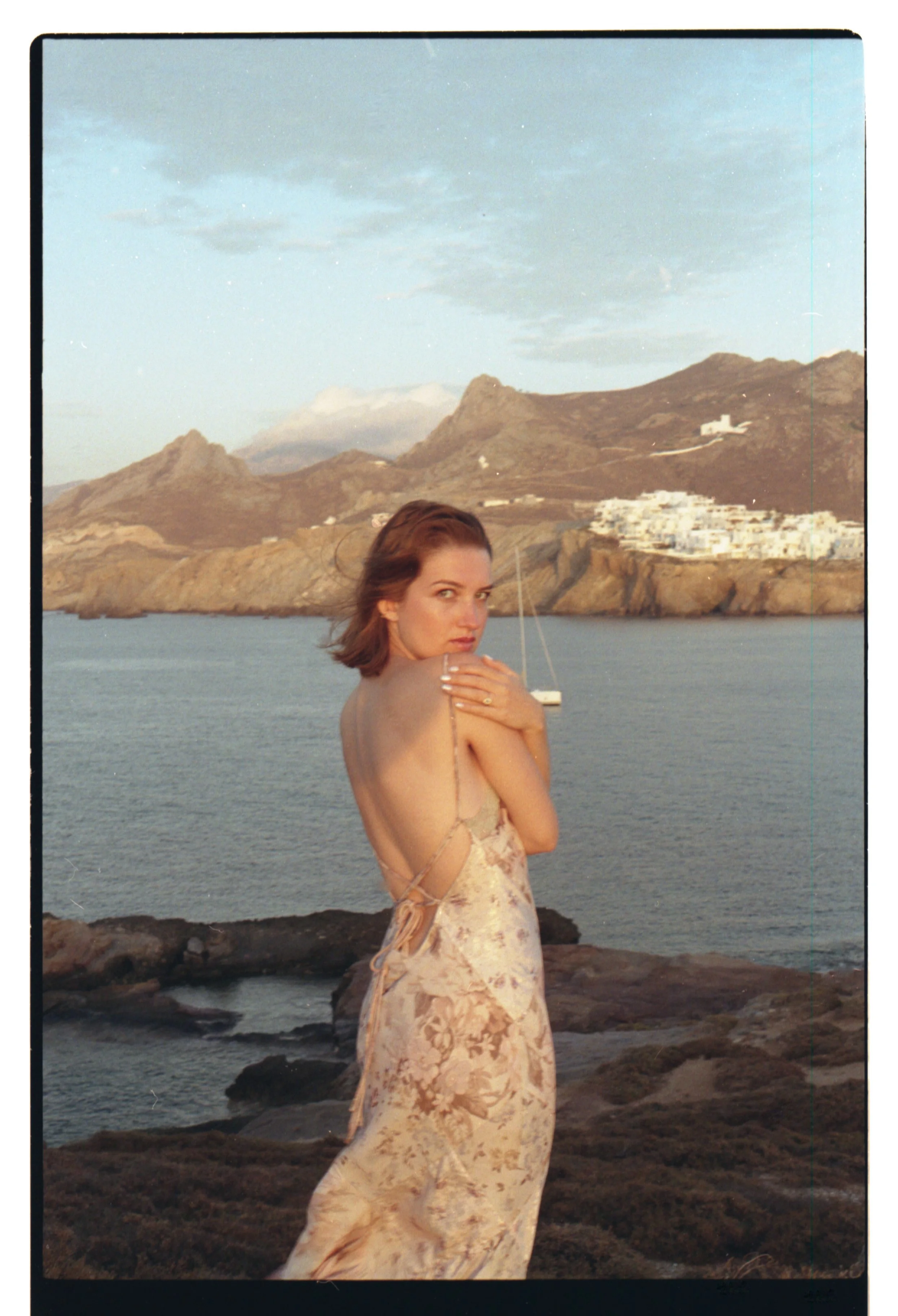 Une femme en robe florale se tourne pour regarder la caméra, avec la mer, des montagnes et un port avec un voilier en arrière-plan.