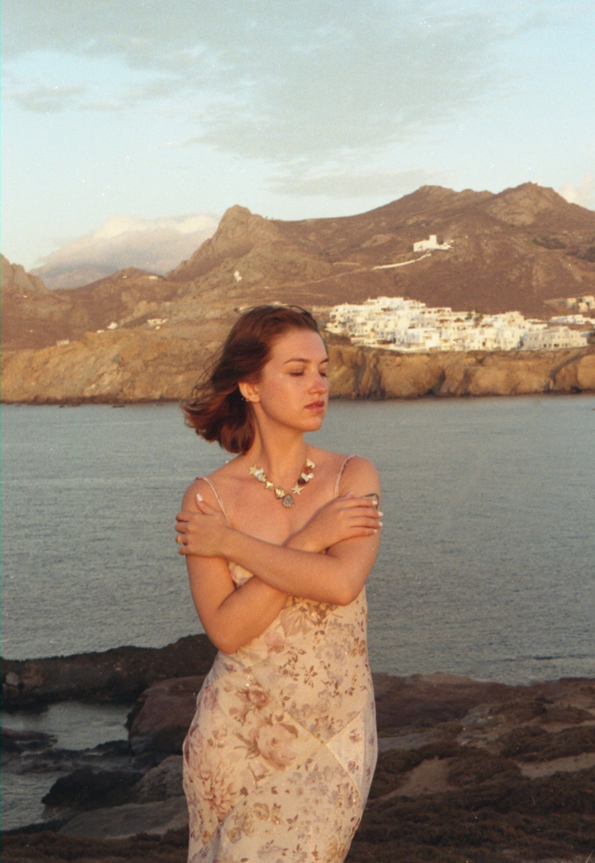 Jeune femme debout près de la mer avec des montagnes en arrière-plan, portant une robe à motifs floraux, un collier en forme d'étoiles, les bras croisés sur la poitrine, avec une expression calme et contemplative.