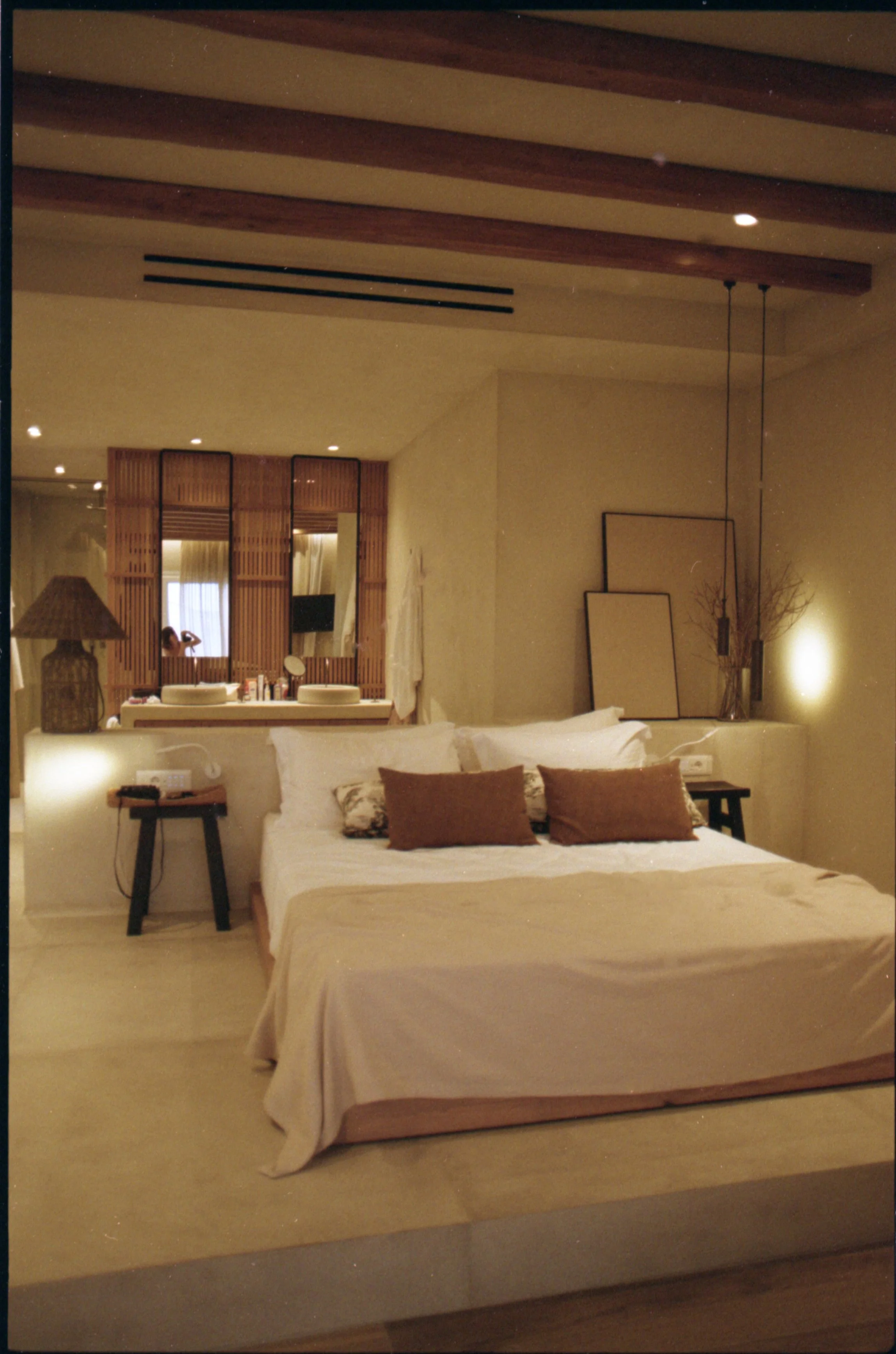 Une chambre d'hôtel élégante avec un lit fait, décorée de coussins marrons et de lampes d'appoint. Un miroir et des cadres vides sont placés contre le mur, avec une lampe sur la table de nuit à gauche. La pièce a un style moderne et chaleureux avec d