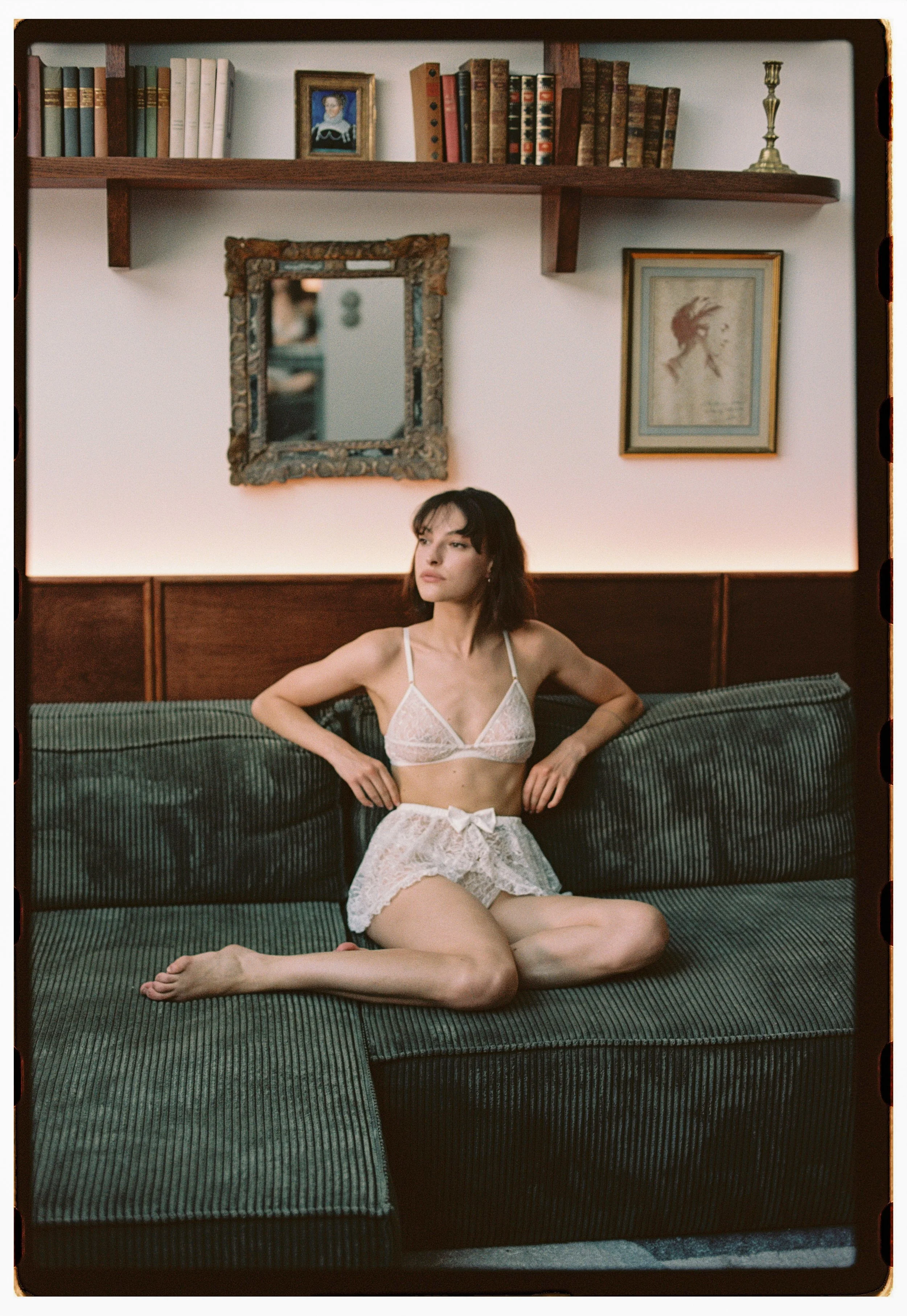 Jeune femme en lingerie blanche assise sur un canapé vert à motifs, dans un salon avec des livres et des affiches anciennes sur le mur.
