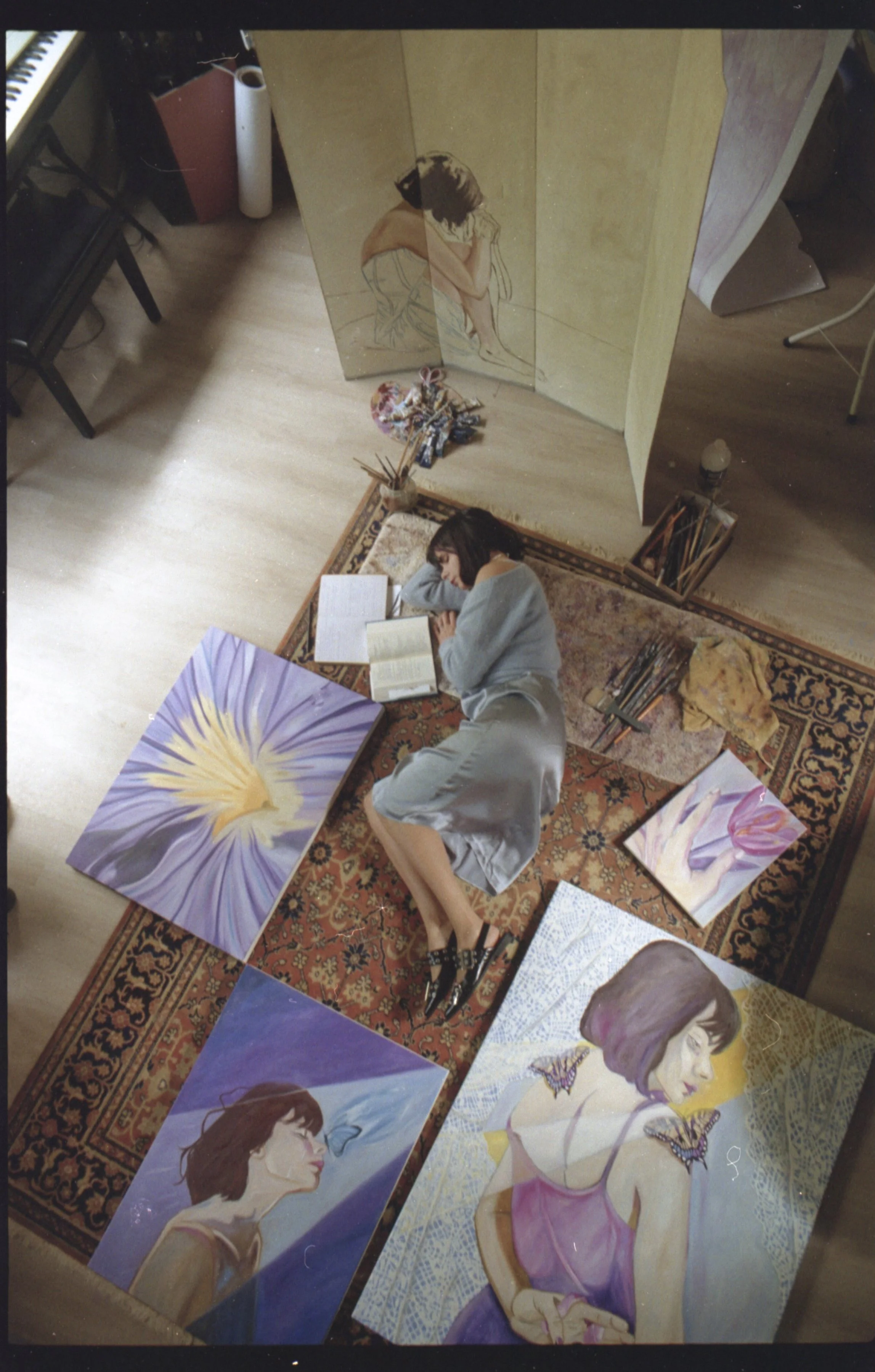Une femme dort sur un tapis, entourée de plusieurs tableaux qu'elle a peints, représentant principalement des portraits de femmes et des fleurs.
