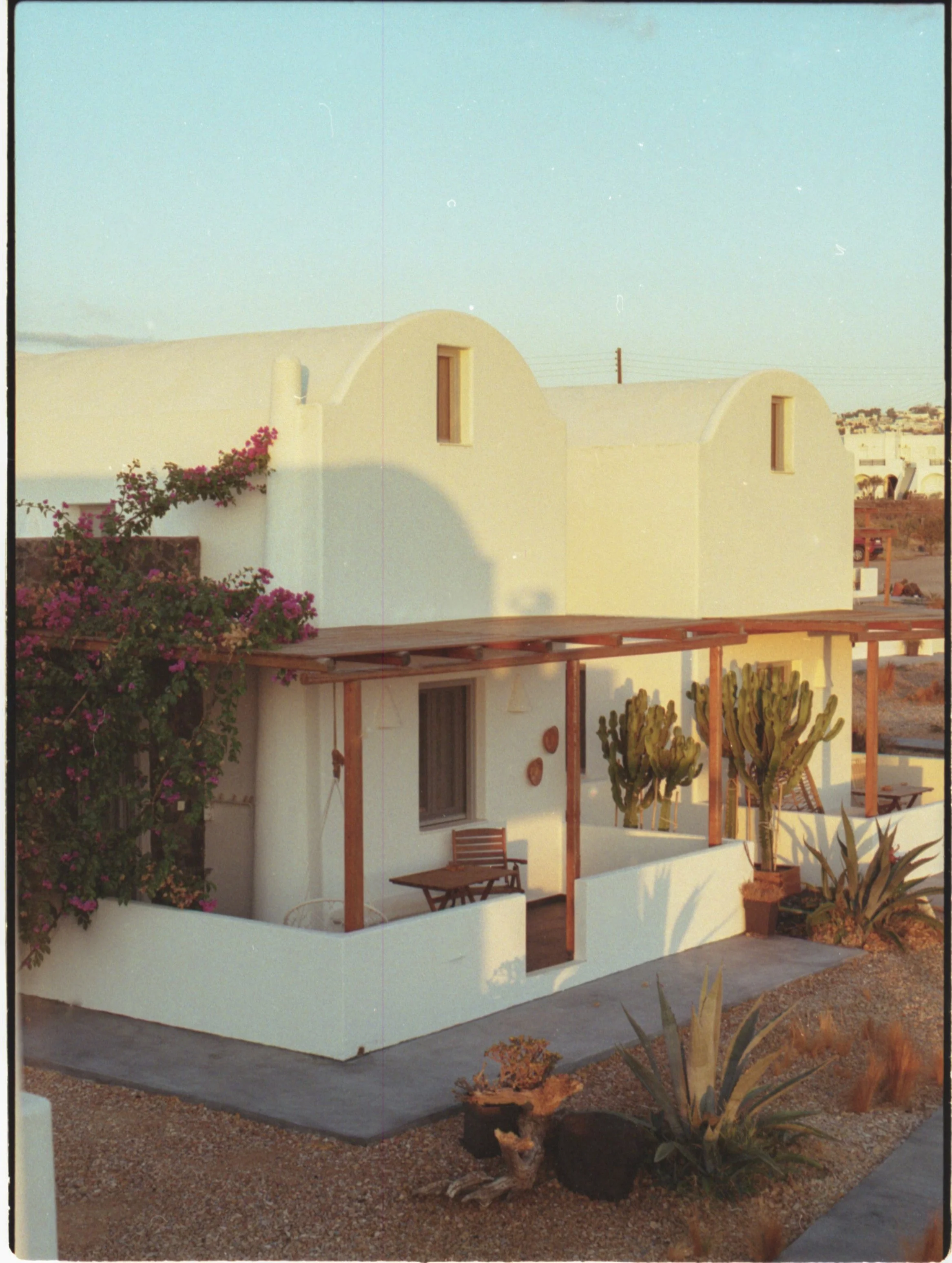 Une maison blanche de style méditerranéen avec des murs courbes, un balcon en bois, des cactus et des plantes dans un paysage désertique au coucher du soleil.