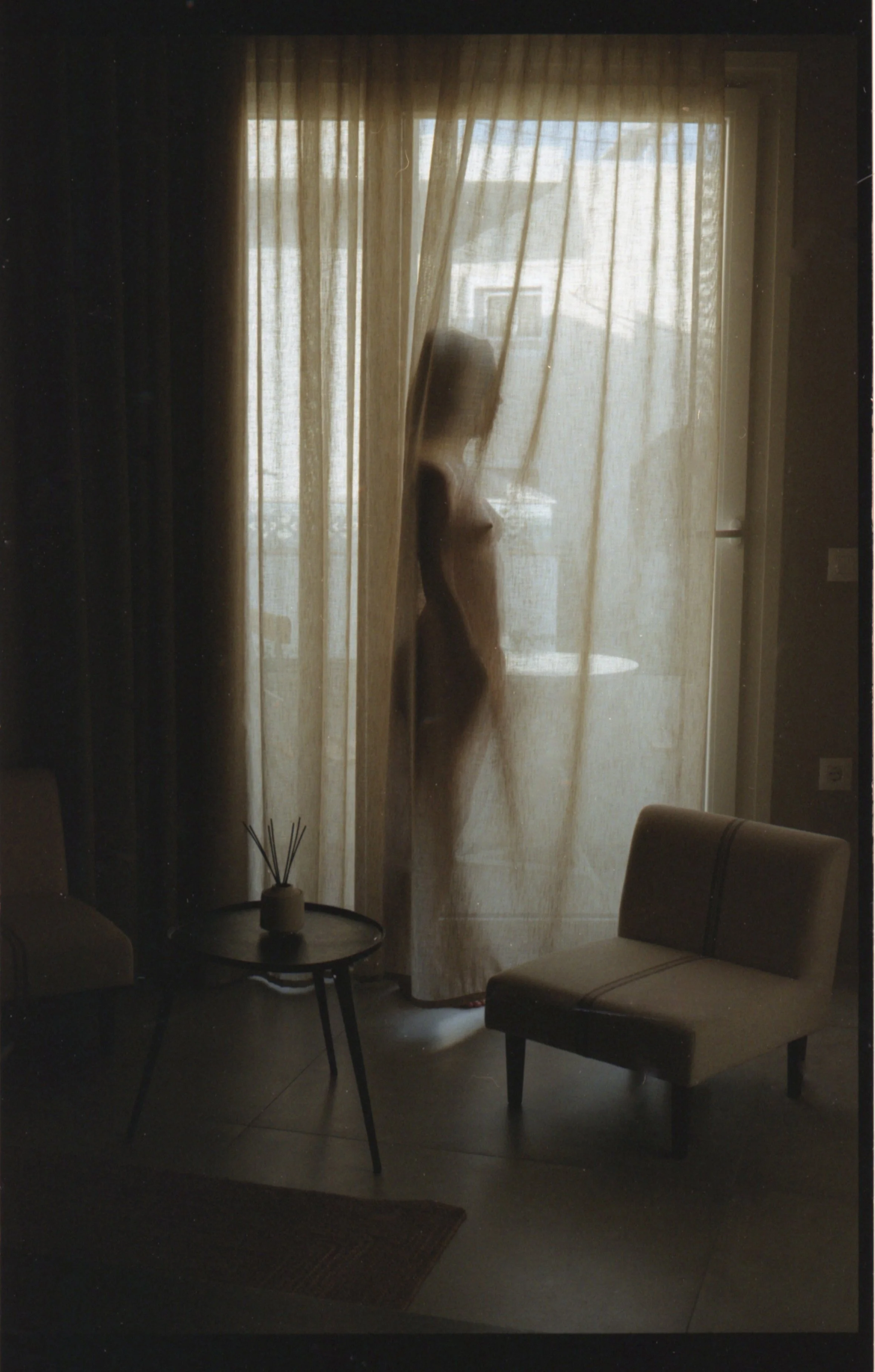 Une silhouette de femme nue derrière des rideaux translucides dans une pièce sombre, avec un canapé et une table à côté