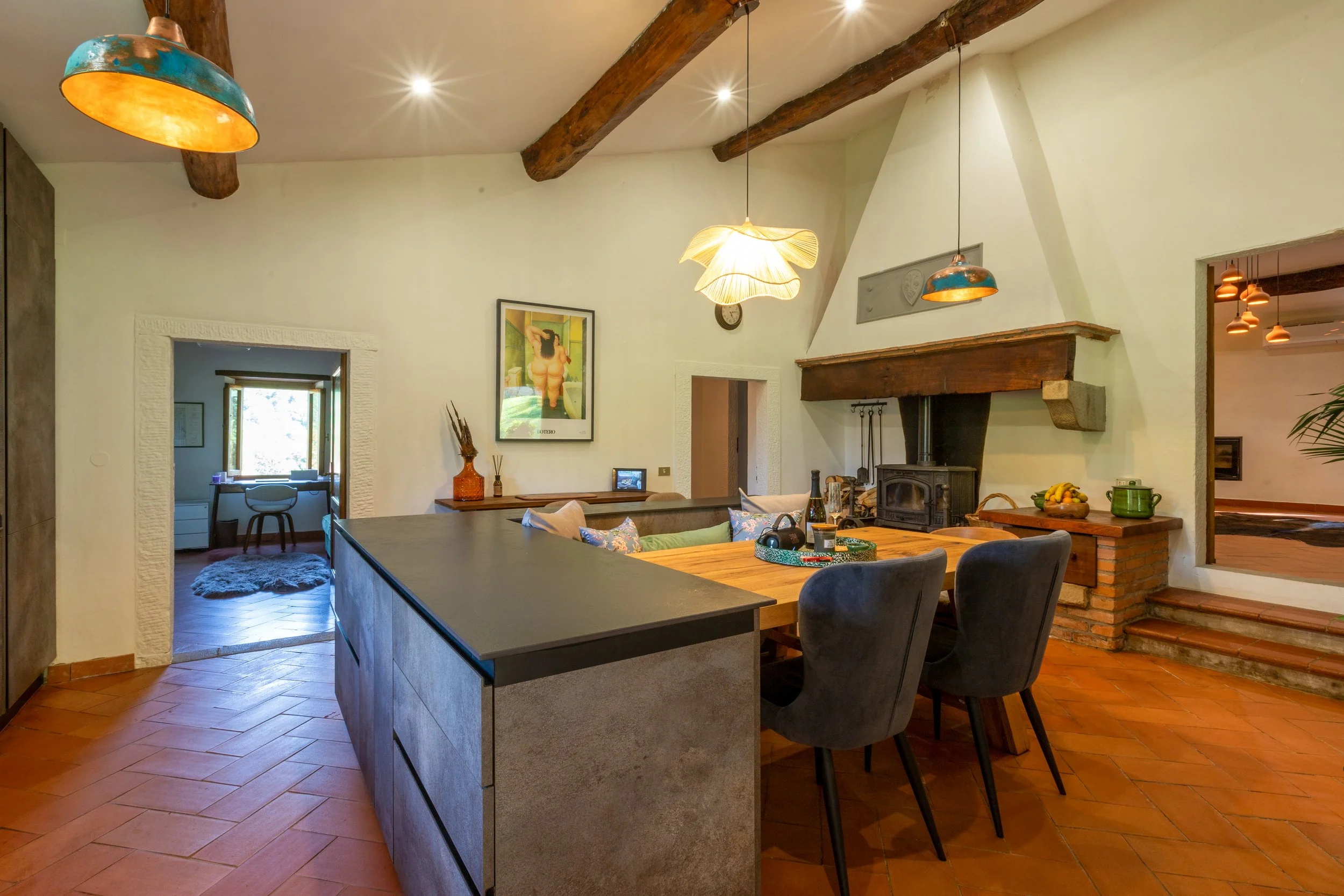 A cozy living room and kitchen area with wooden accents, a wood stove, a large dining table with chairs, and decorative lighting fixtures hanging from the ceiling.
