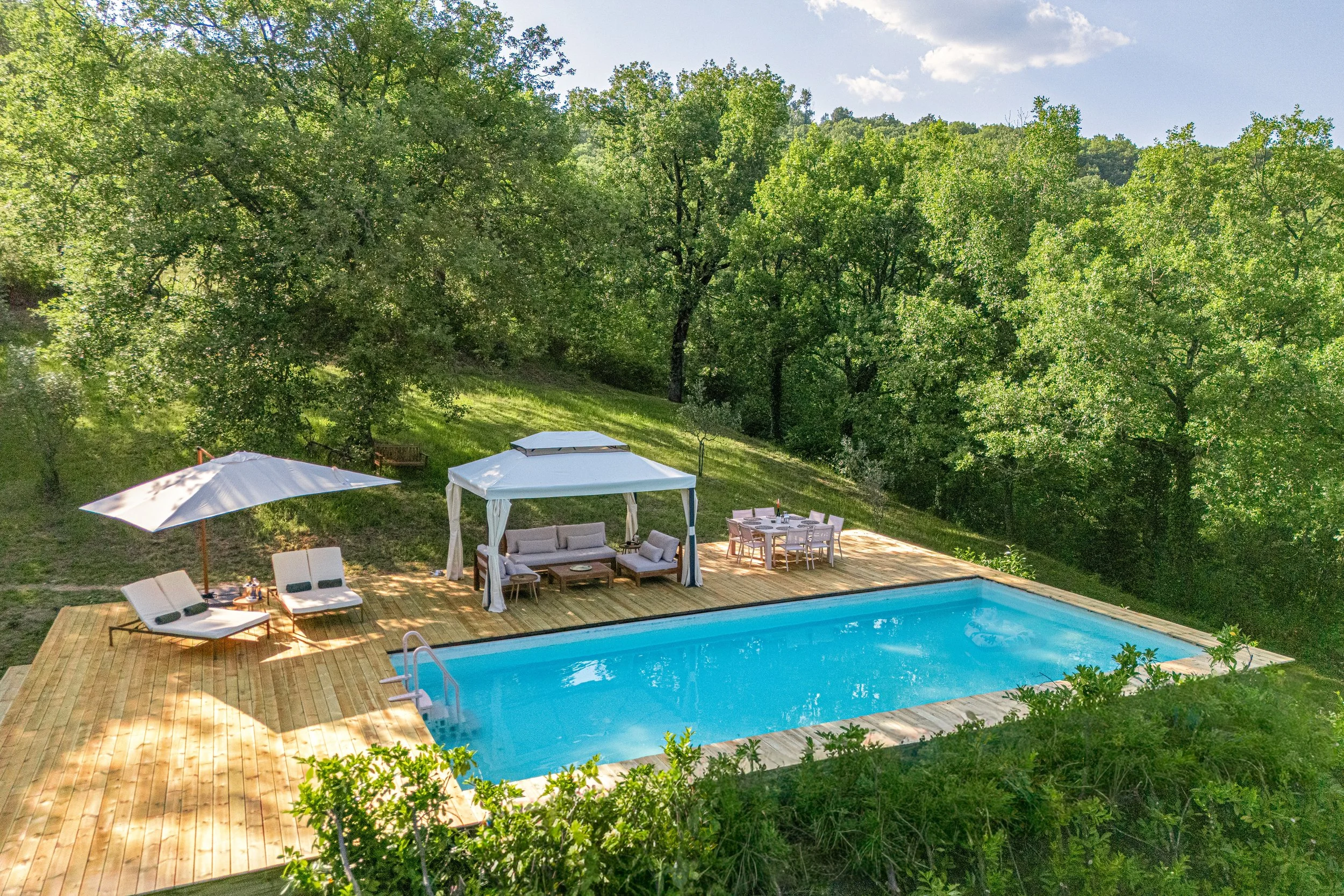 A backyard scene with a swimming pool, outdoor furniture including lounge chairs, a shaded seating area, and surrounding trees under a partly cloudy sky.
