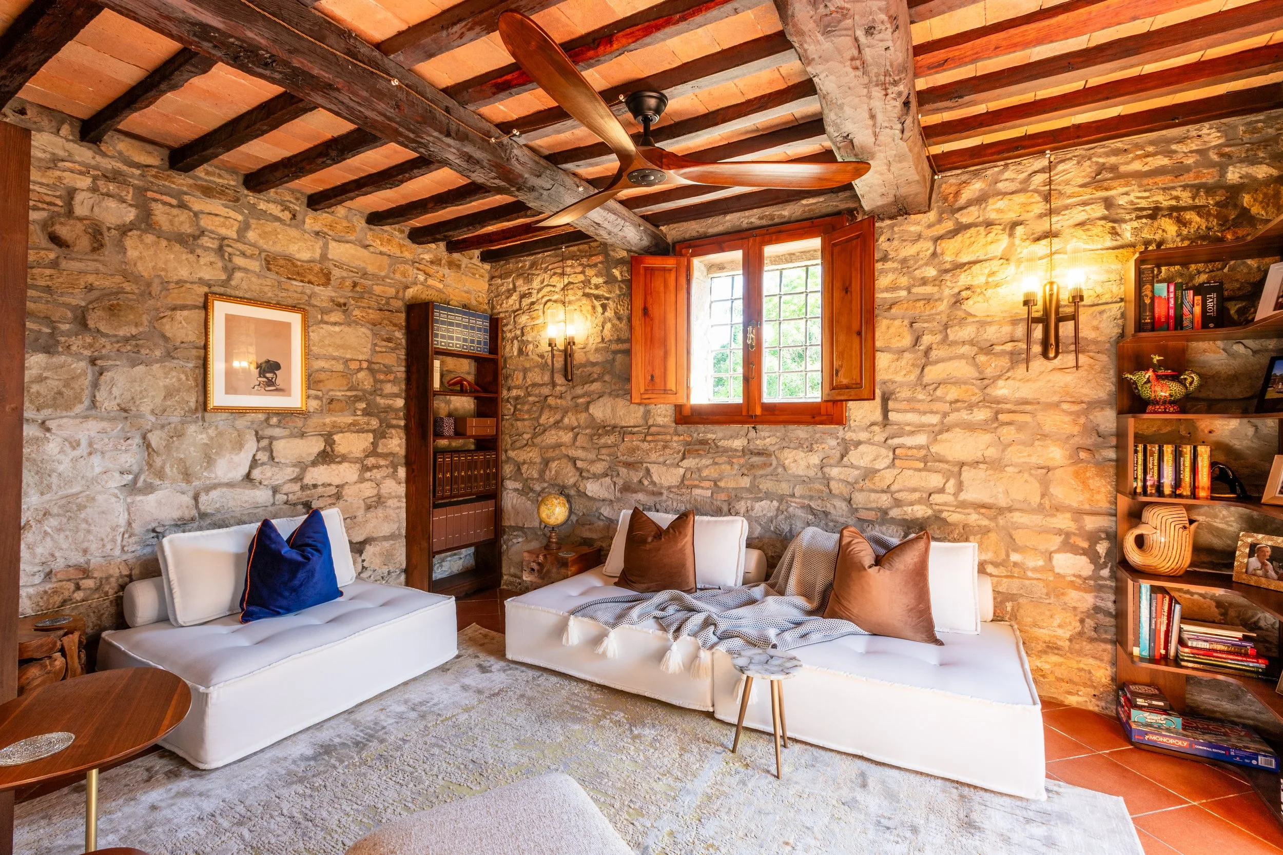 A living room with exposed stone walls, wooden ceiling beams, and a ceiling fan. There are two white sofas with colorful pillows, a small wooden side table, a bookshelf, and a window with wooden shutters letting in natural light. Decor items include framed artwork, a globe, and a lamp with multiple bulbs.