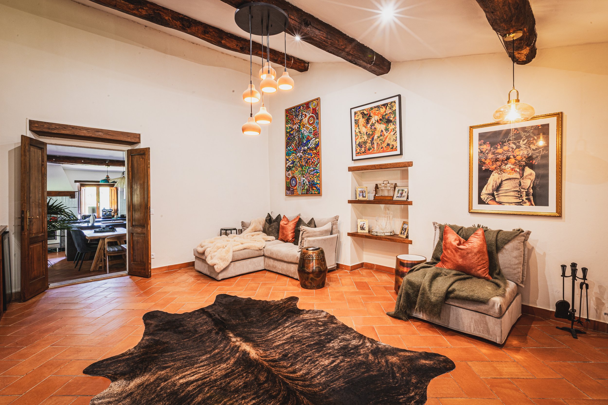 A living room with terracotta tile flooring, beige couches with pillows and blankets, wall art, and a black and brown cowhide rug in foreground. Open doorway reveals a dining area with wooden table and chairs, and potted plant. Exposed wooden beams on ceiling and framed pictures on wall.