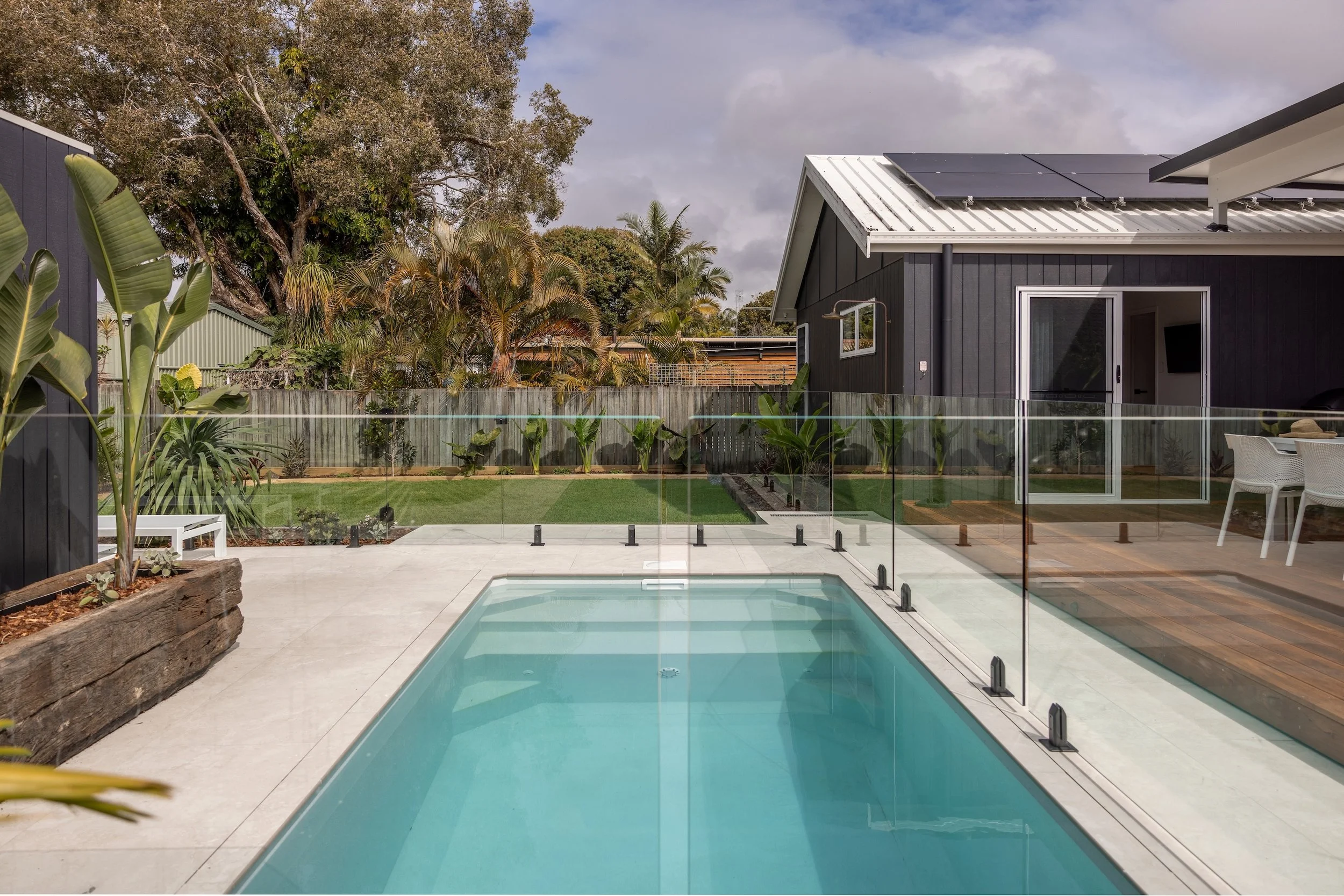 Backyard with small pool enclosed by glass fencing, surrounded by plants and trees, adjacent to a modern black house with solar panels on the roof.