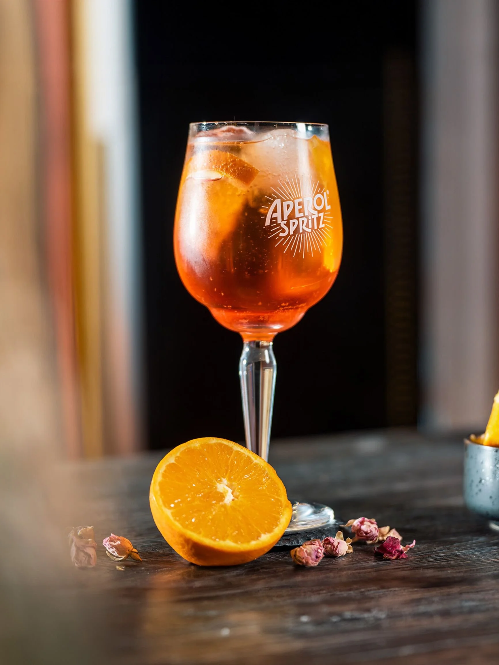 Ein Glas Aperol Spritz mit Eis und Orangenscheiben auf einem Holztisch, daneben eine halbe Orangenscheibe und getrocknete Rosenknospen.