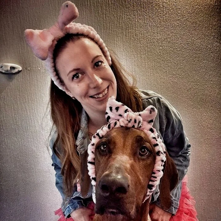 Eine Frau und ein Hund, beide tragen pinke, gepunktete Tierohren-Accessoires. Die Frau trägt einen passenden Haarreif und hat braune Haare, das Tier ist ein brauner Hund.