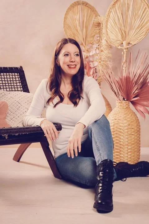 Eine lachende Frau mit braunen Haaren sitzt auf dem Boden vor einem Pendellehnen neben einer großen Vase mit getrockneten Blumen.