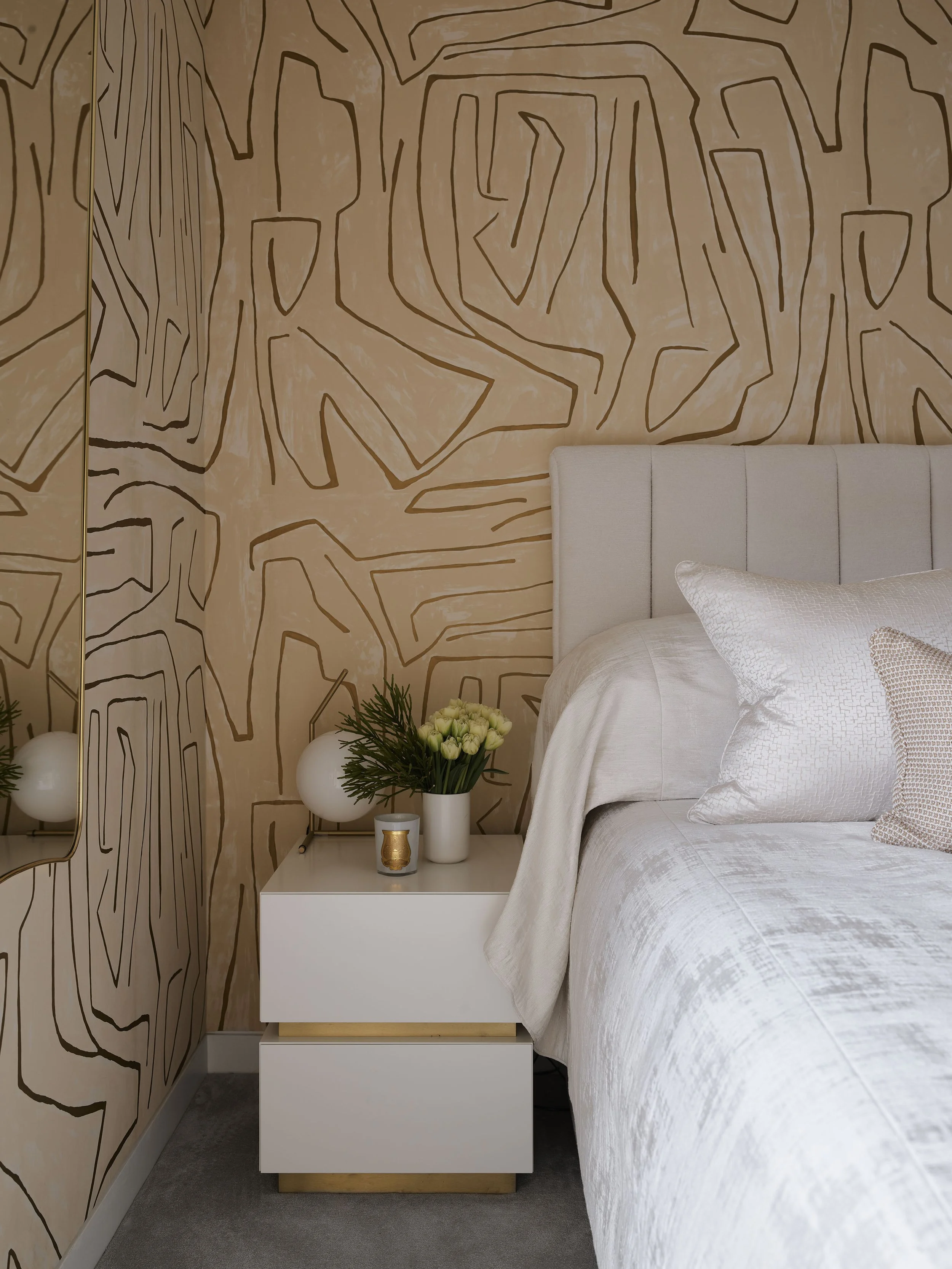 A modern bedroom corner with a white upholstered headboard, white bedding, and pillows. A white bedside table holds a vase with white roses and greenery, a small candle, and two spherical lamps. The wall has an abstract, geometric pattern in neutral 