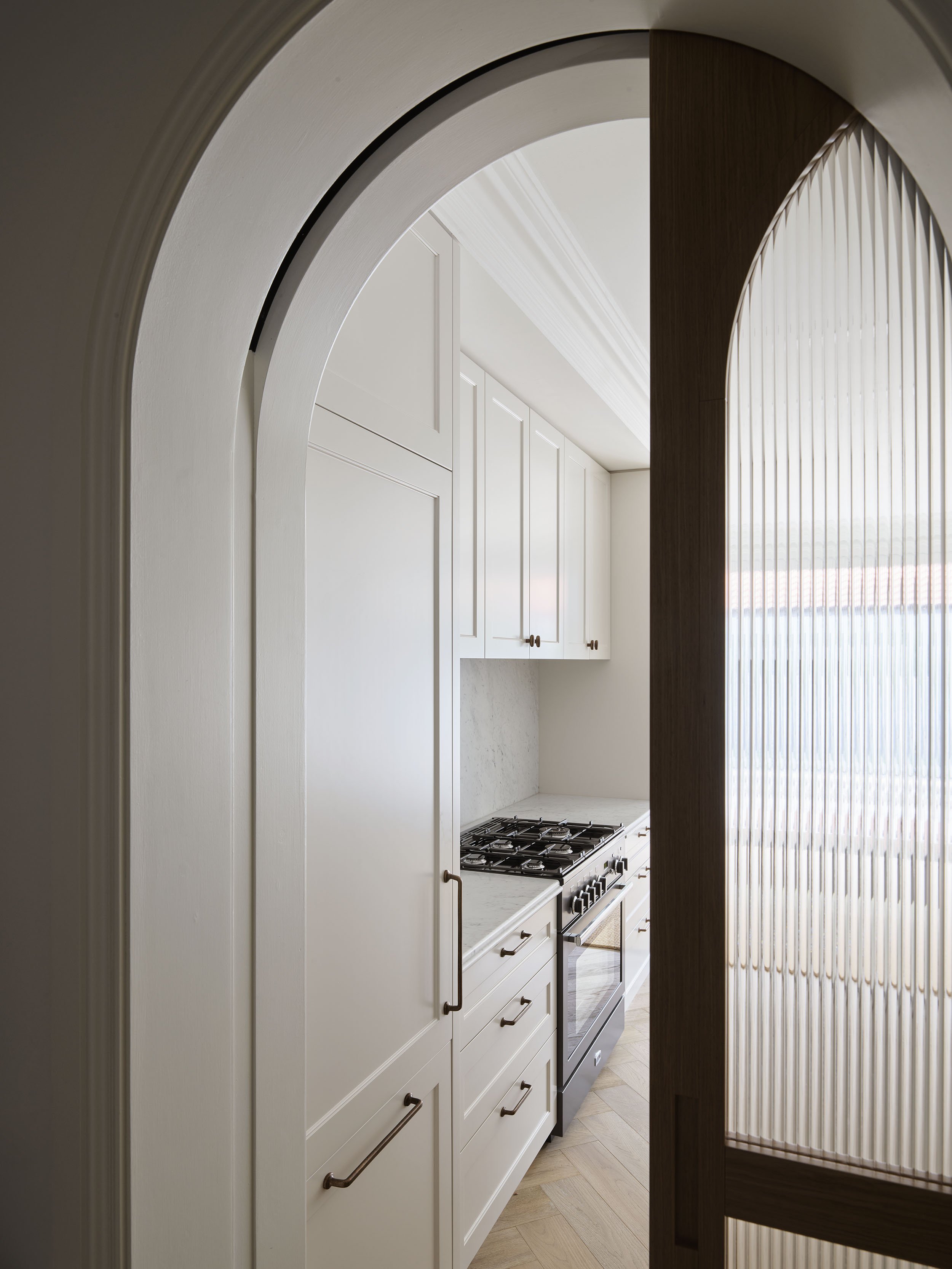 View through an arched doorway into a modern kitchen with white cabinets, marble countertops, and a stainless steel stove, with textured glass door on the right side.