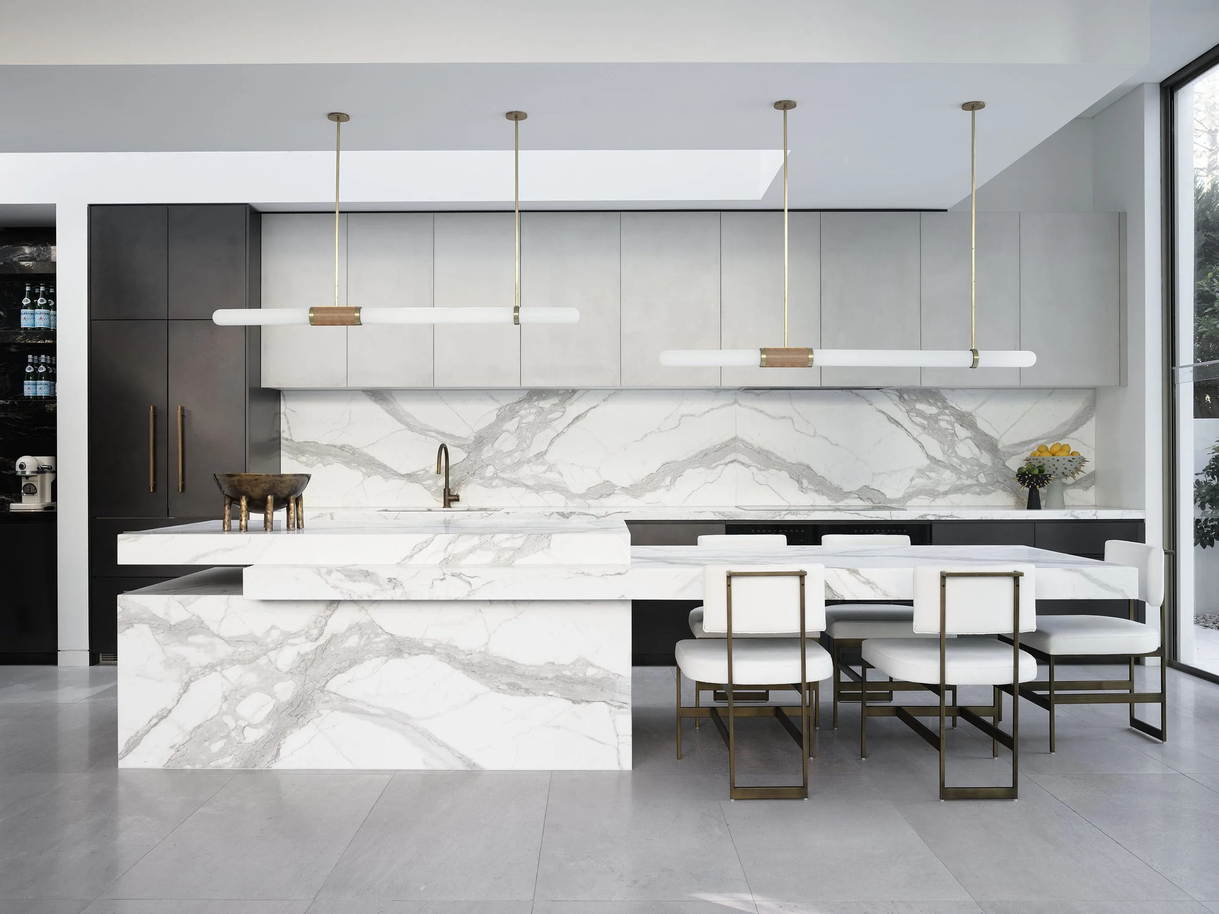 Modern kitchen with marble island, dark and light cabinetry, pendant lights, and white chairs with gold frames.