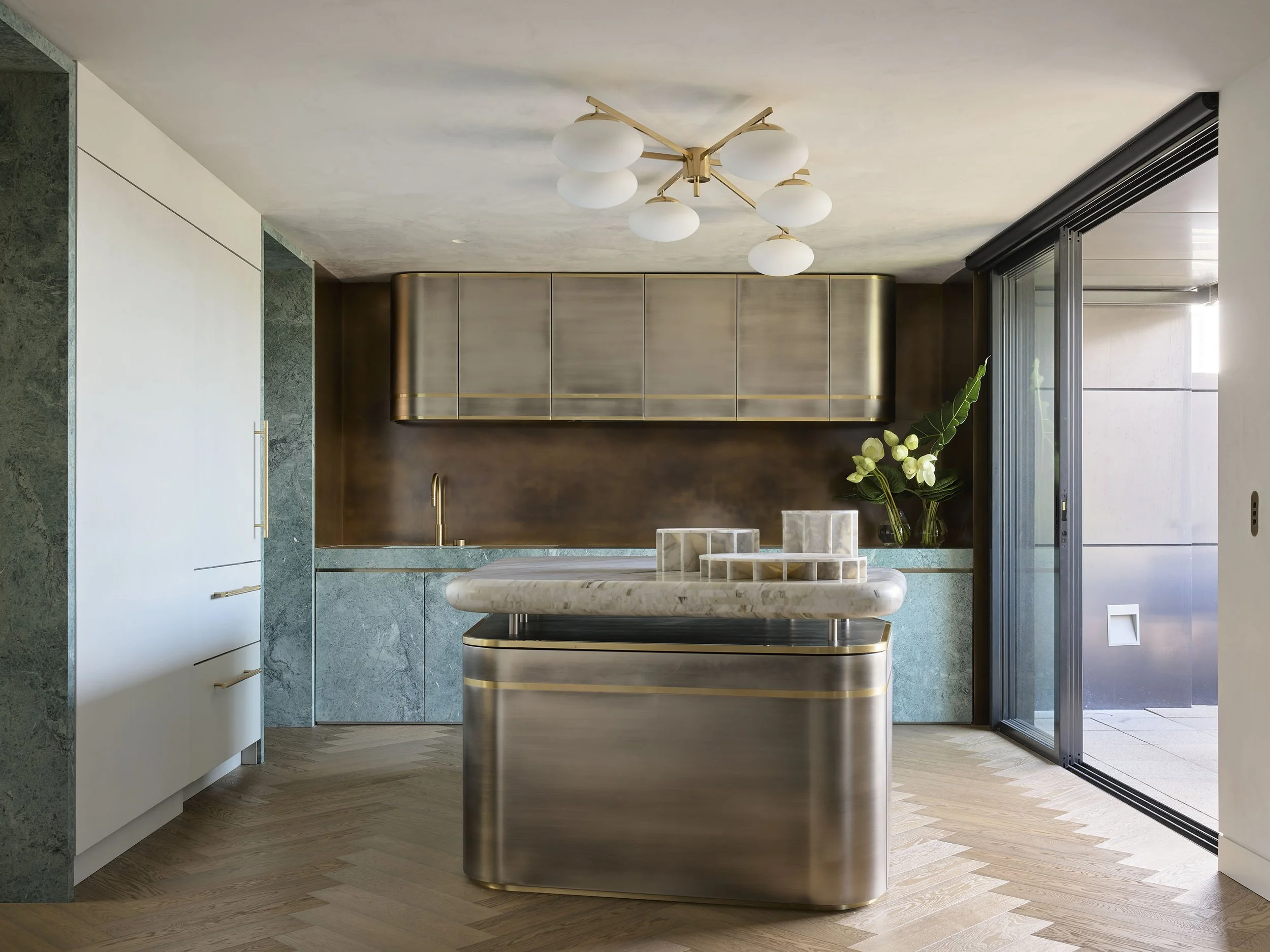 Modern kitchen with marble island, brass cabinetry, green marble countertops, and a large sliding glass door leading outside. Green plant with white flowers on the countertop.