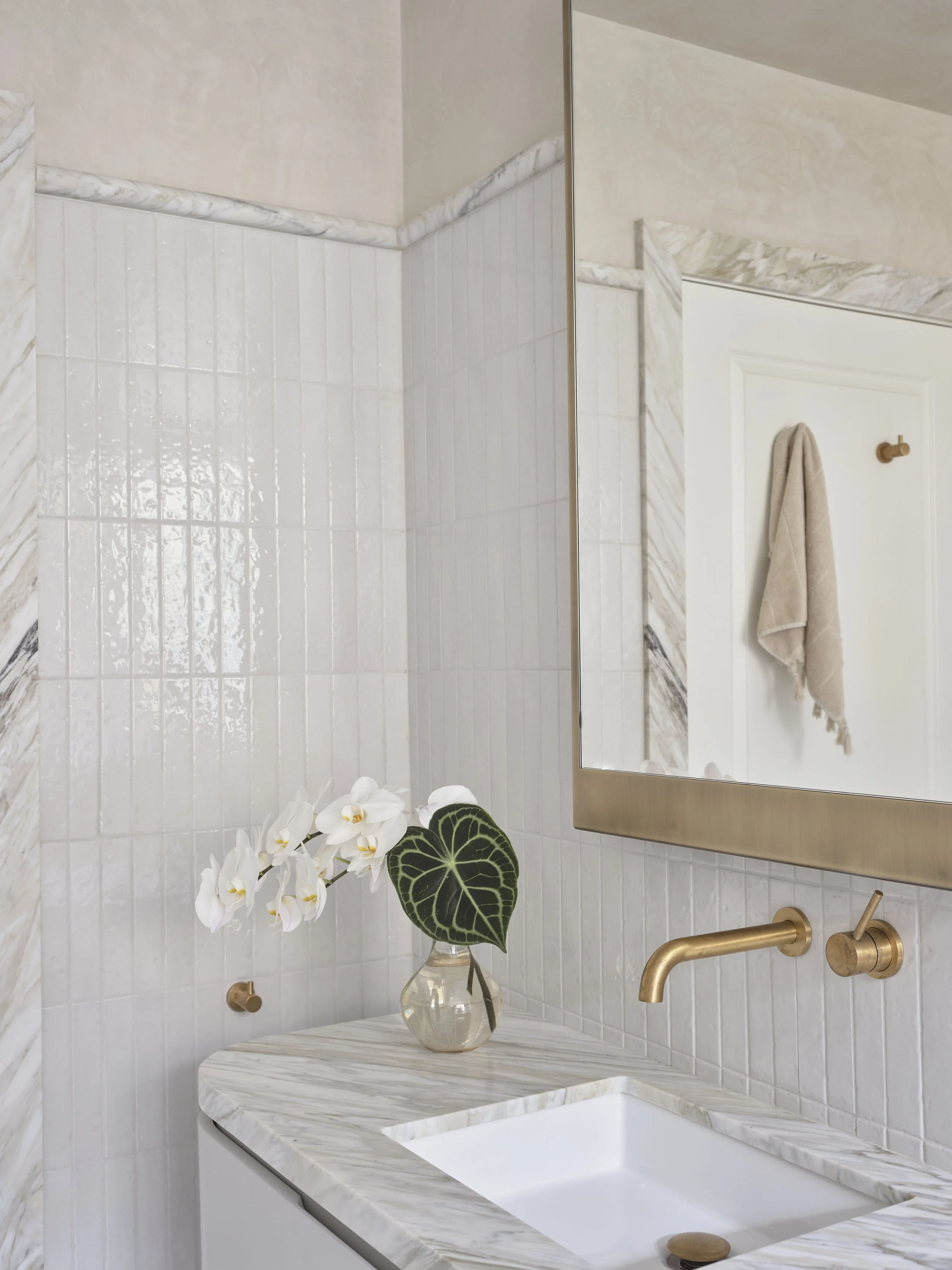 Bathroom with white marble countertop, a white sink, and a mirror with a gold frame. There is a vase with white orchids and a large green leaf. A beige towel hangs on the wall.